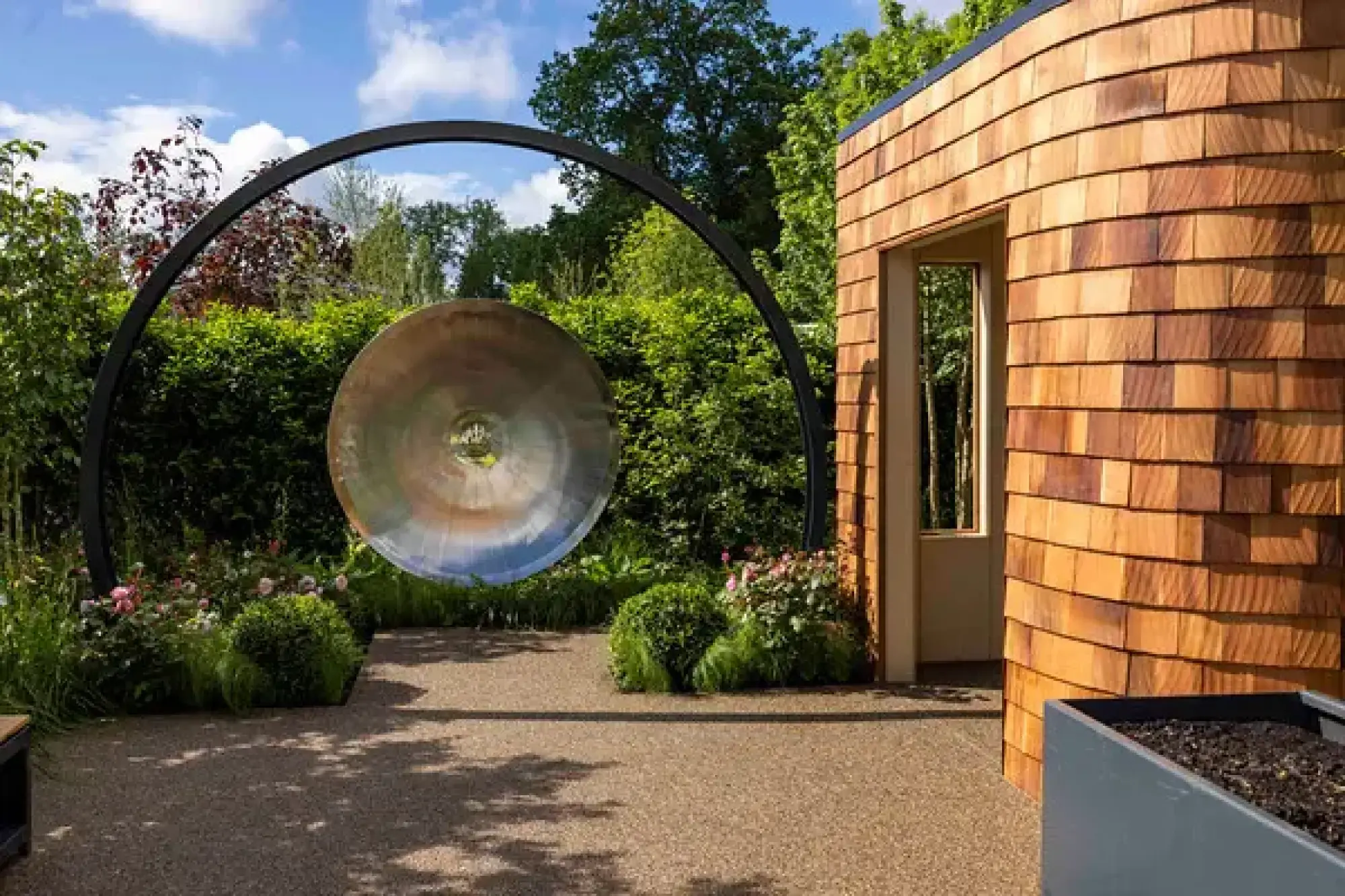 A large metallic gong within a black arch stands on an aggregate path, beside a curved building clad in timber shingles and surrounded by green shrubs and pink roses.