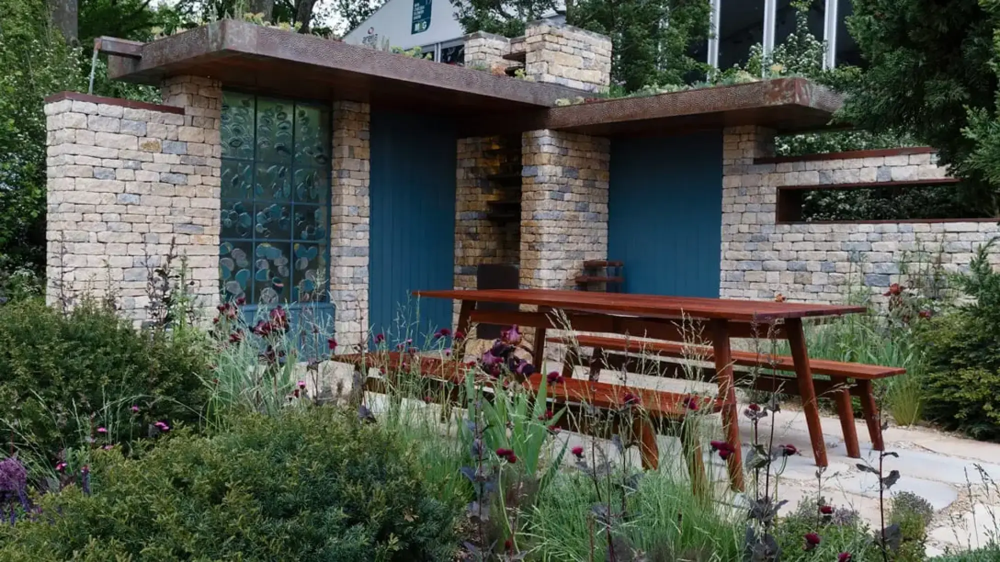A show garden features a dry stone building with a Corten steel roof, stained glass, teal panels, and a timber picnic table.