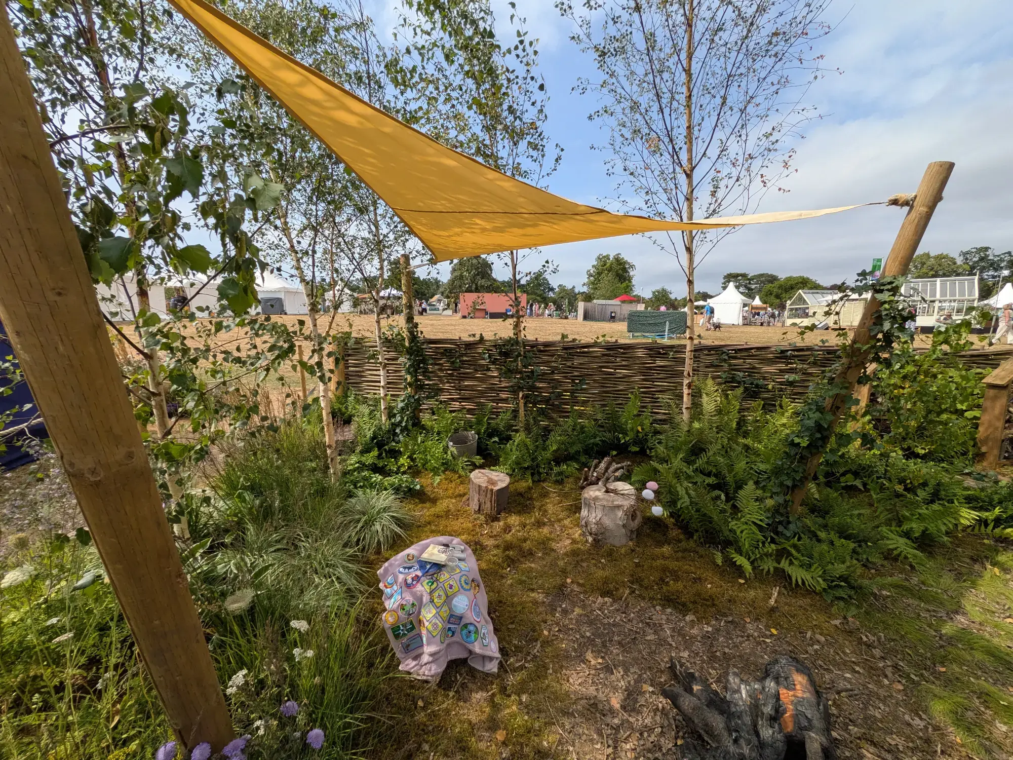A rustic woodland camp garden features a yellow shade sail, log stump seating, a campfire area, silver birch trees, ferns, and a badge-covered blanket on mossy ground.