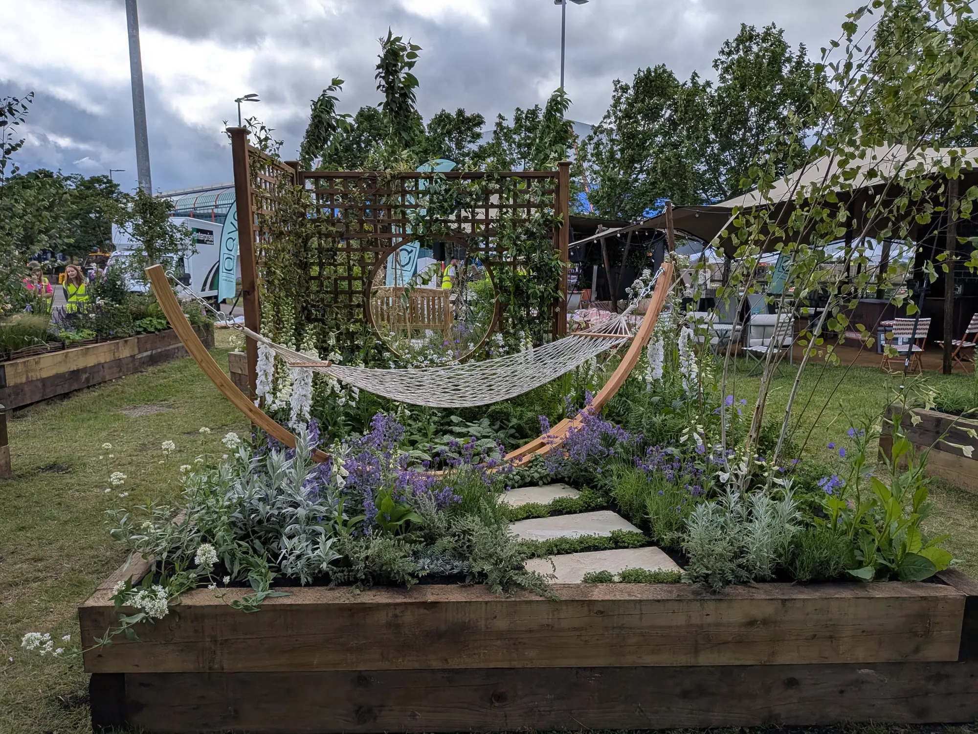 A tranquil garden border with a white rope hammock, a circular mirror in a wooden trellis, and a path through white and purple flowers.