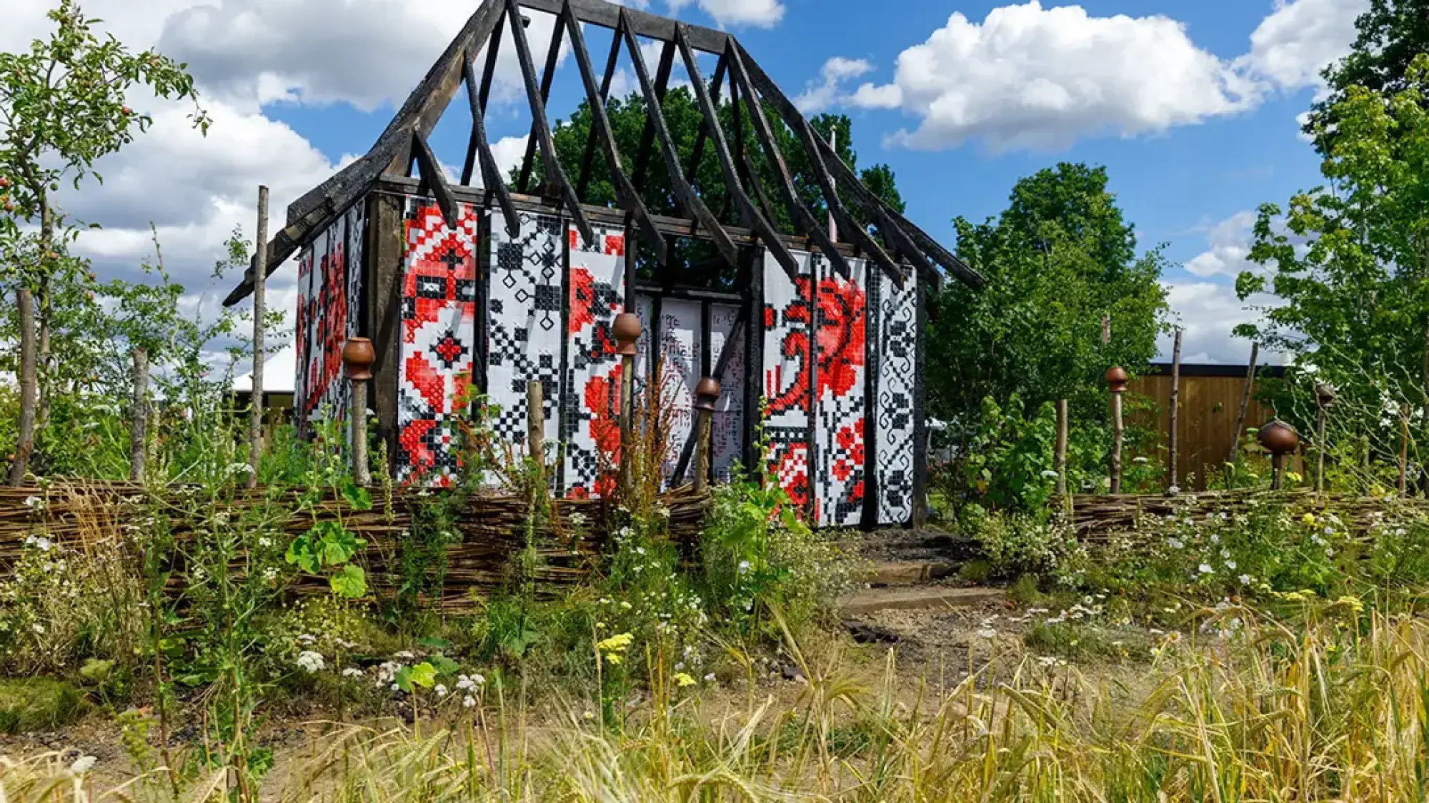 A garden featuring a burnt timber house with mosaic walls in red, black, and white Ukrainian patterns, surrounded by a wattle fence and wild meadow planting.