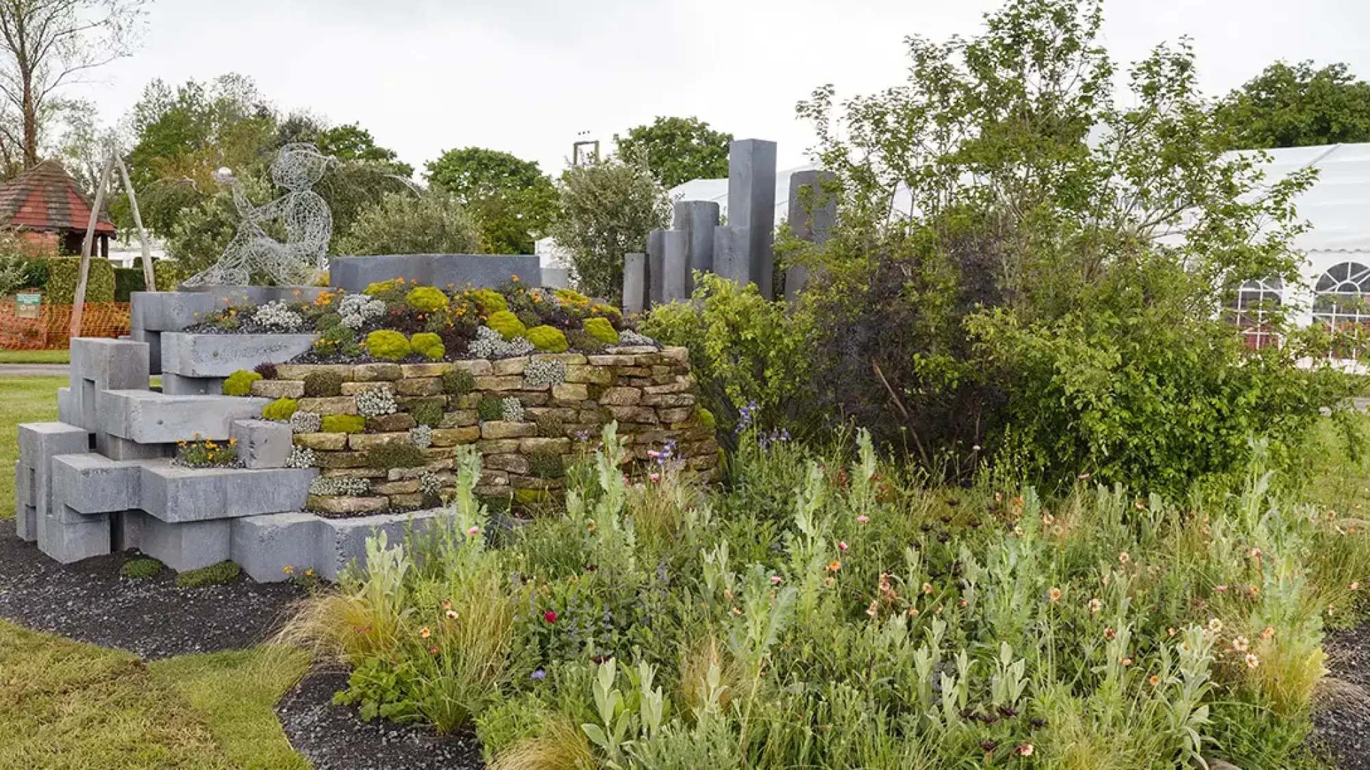 A modern show garden features a wireframe sculpture of a seated figure, stepped grey block walls, and a dry stone wall, with lush succulent and meadow planting.