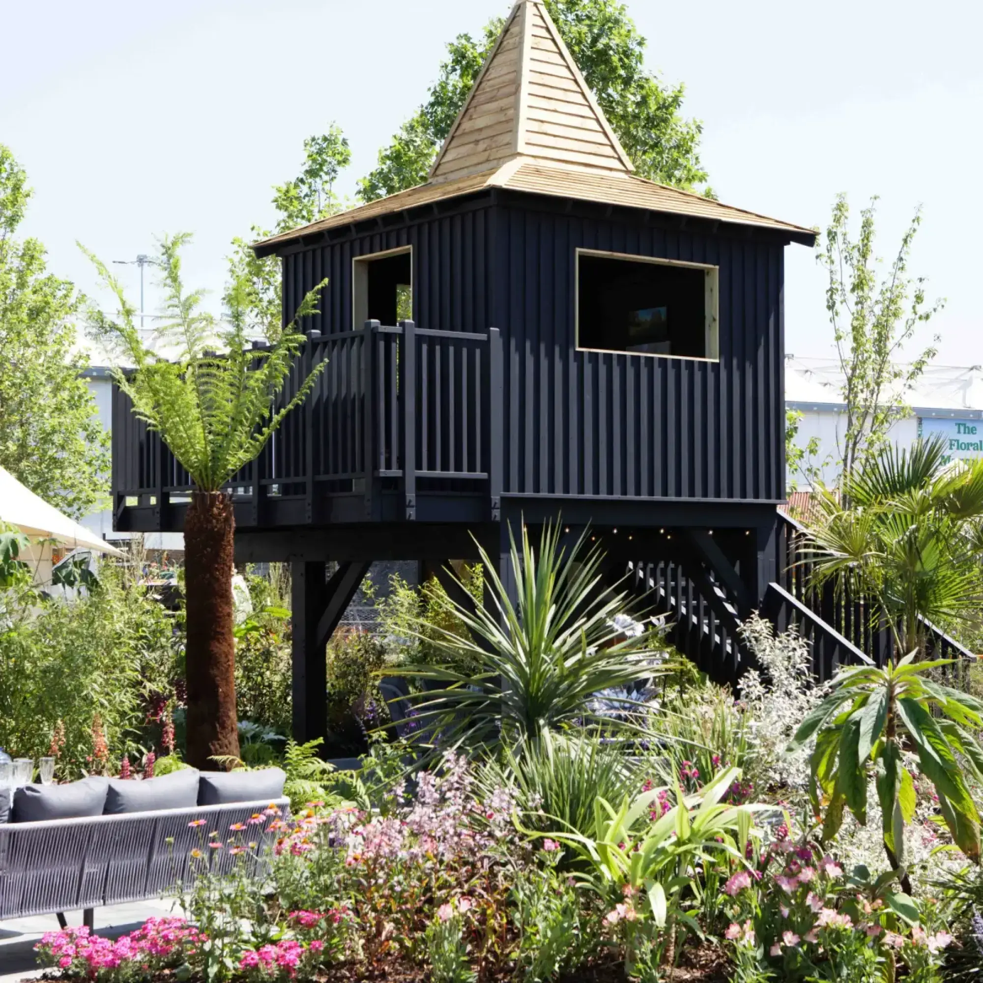 An elevated black timber cabin with a pointed wooden shingle roof is surrounded by lush green foliage, vibrant pink and purple flowering plants, and a grey outdoor sofa.