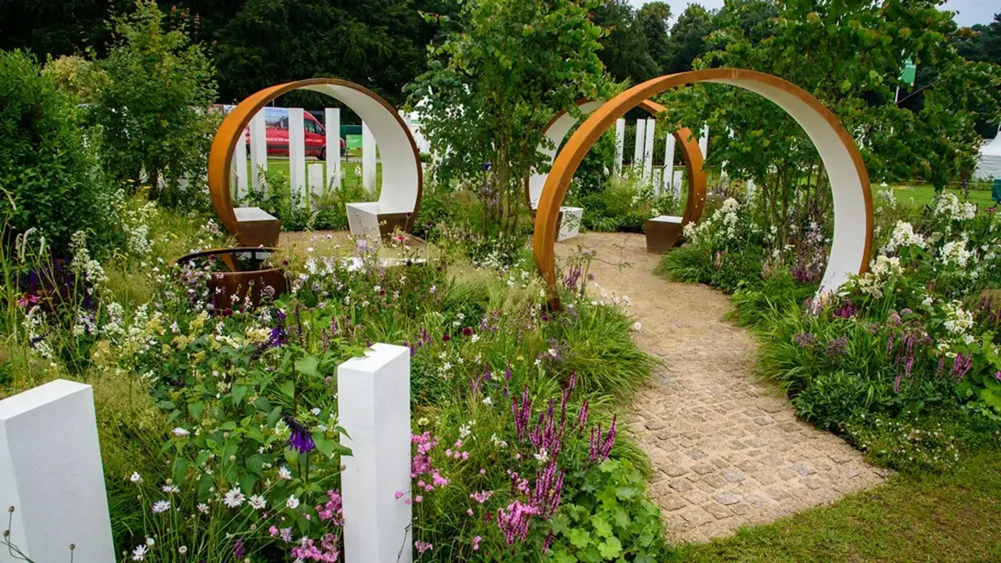 A modern garden with two large rusted steel arches, each with a white bench, flanking a cobbled path surrounded by lush white and purple perennial planting, a small water feature, and white pillars.