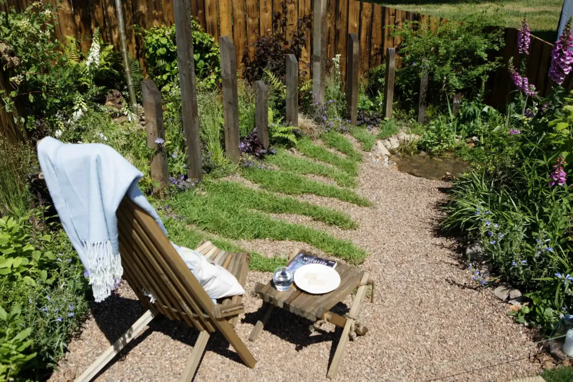 A naturalistic garden features a gravel path with grass stripes, a small pond, and a wooden chair and table among lush green and purple planting.