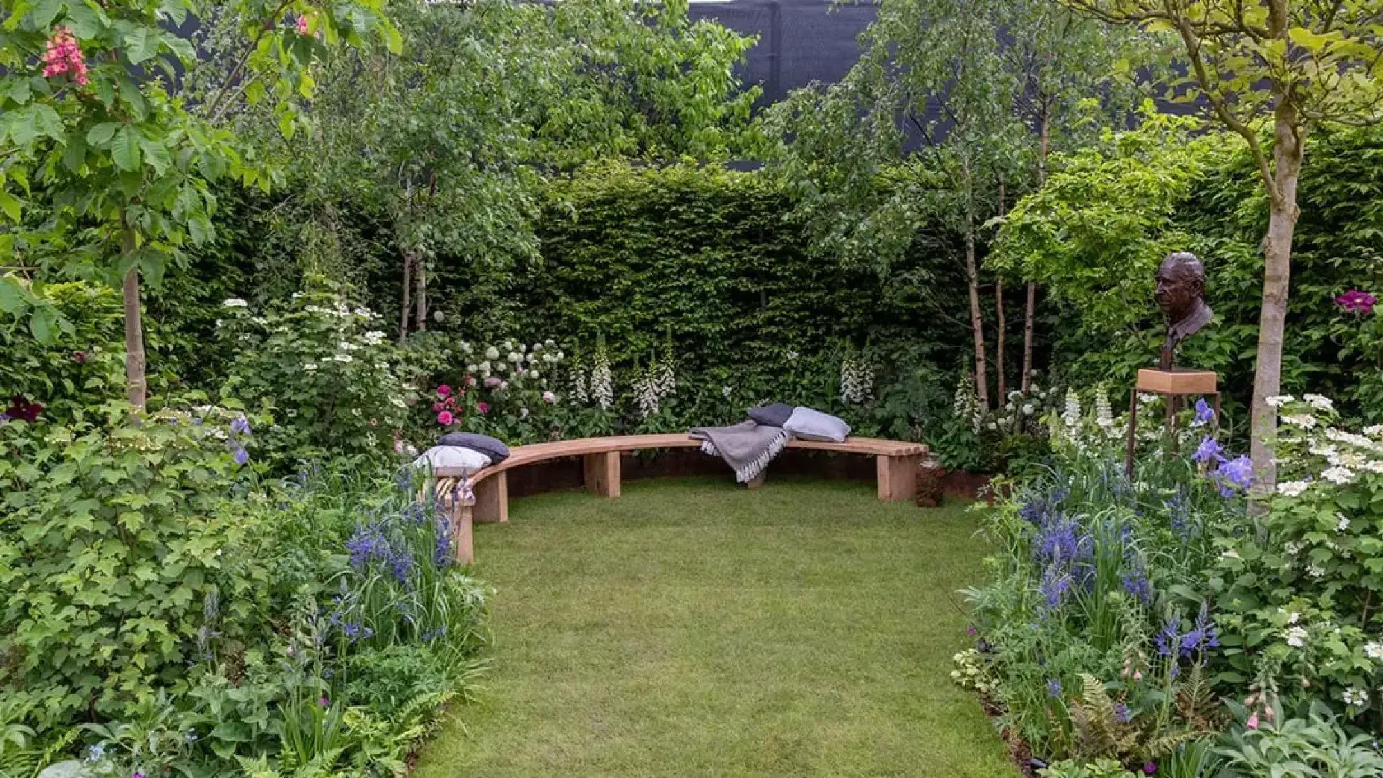 A tranquil feature garden with a bronze bust sculpture, a long curved timber bench on a lawn, surrounded by lush mixed borders of mature trees, flowering shrubs, and perennials in shades of blue, white, and pink.
