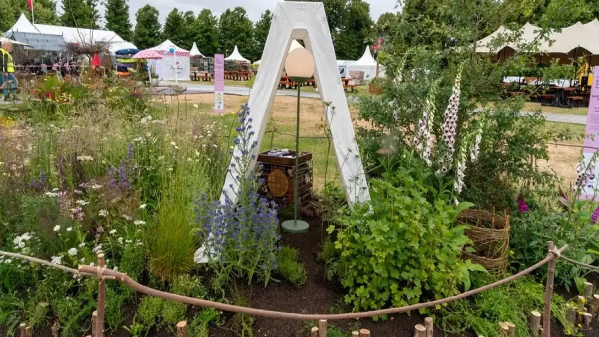 A naturalistic pocket garden featuring a white A-frame structure with a spherical lantern, a moth house, and diverse wildflowers including white and purple foxgloves.