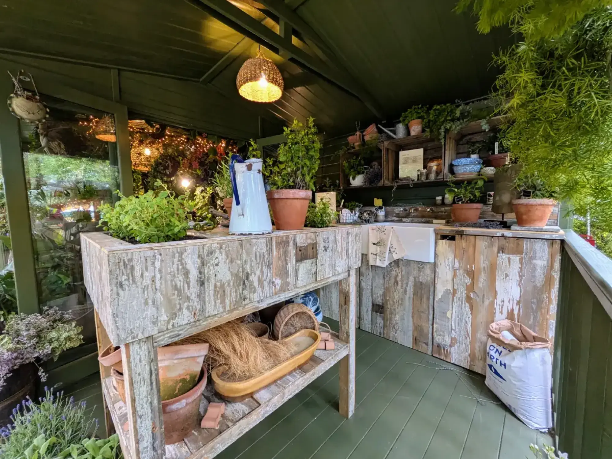A green houseplant studio with a shabby chic reclaimed wood potting bench and shelves displaying potted plants, a white jug, and a wicker light.