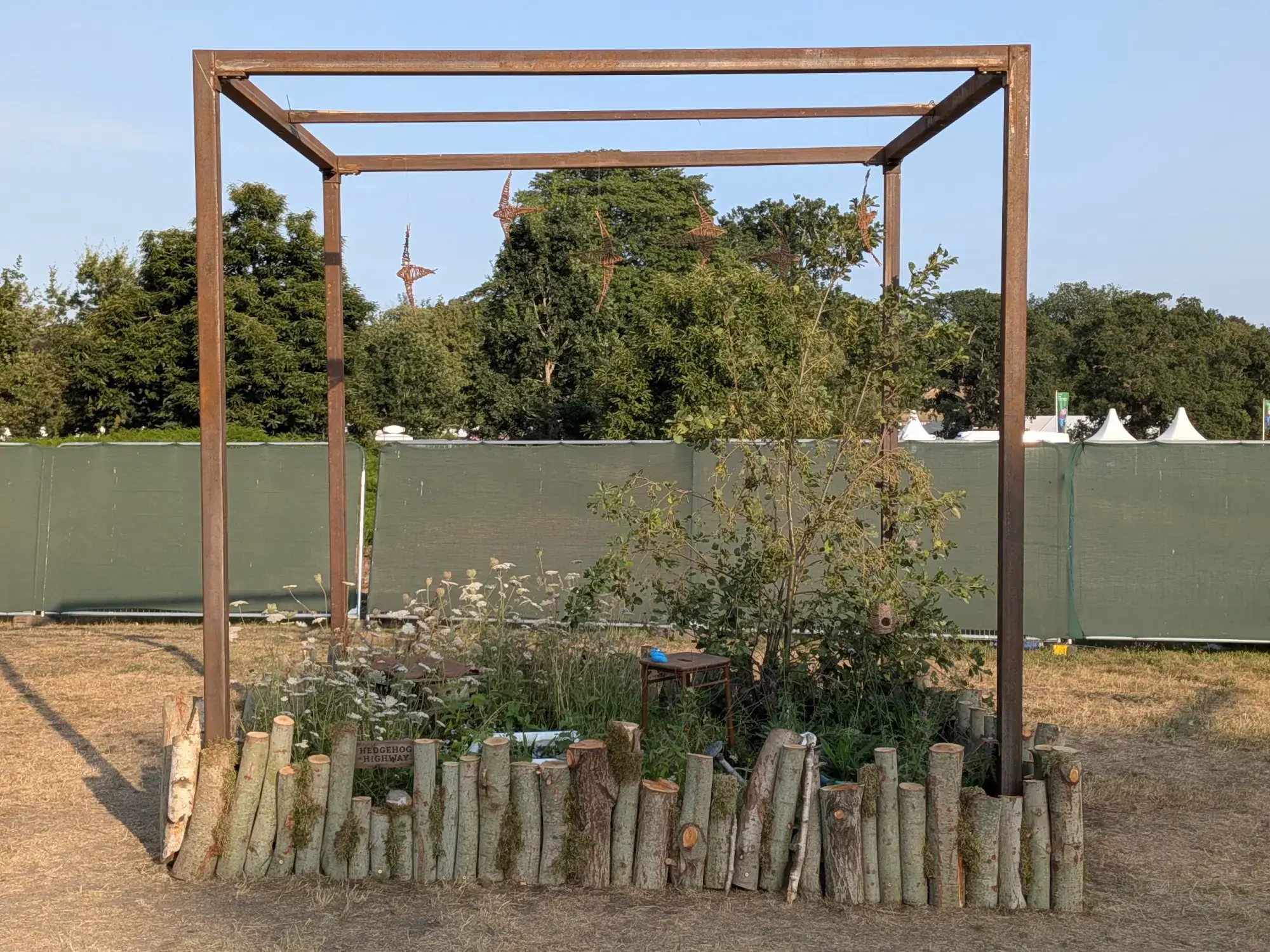 A rustic cube garden featuring a rusted steel frame enclosing naturalistic planting, a log pile border, a small tree, wildflowers, and elements designed for wildlife.