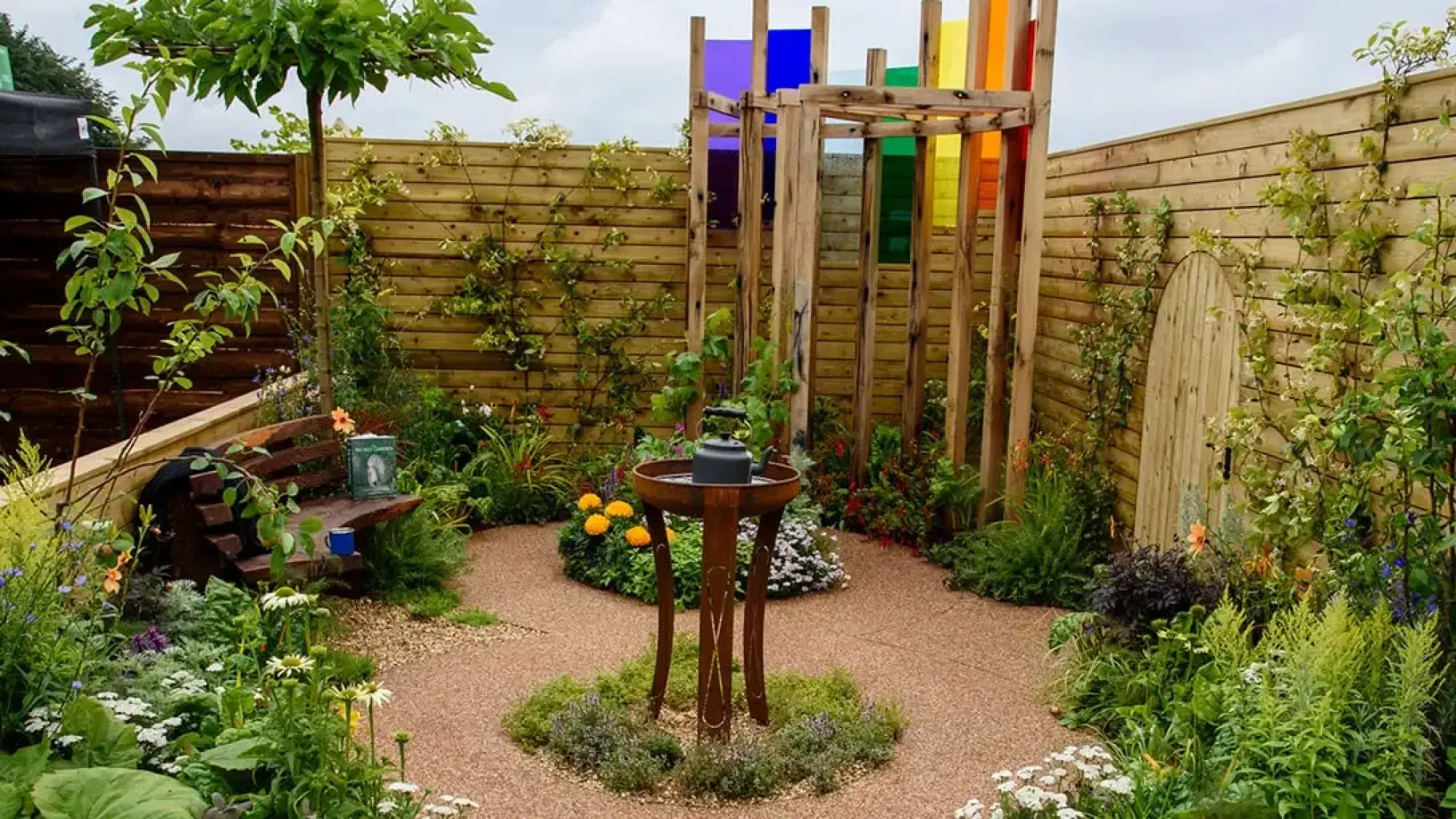 A tranquil terrace garden features a curved gravel path, a wooden pergola with rainbow coloured glass panels, and a rusted metal bird bath, surrounded by lush, informal planting and a timber fence with a hidden wooden door.
