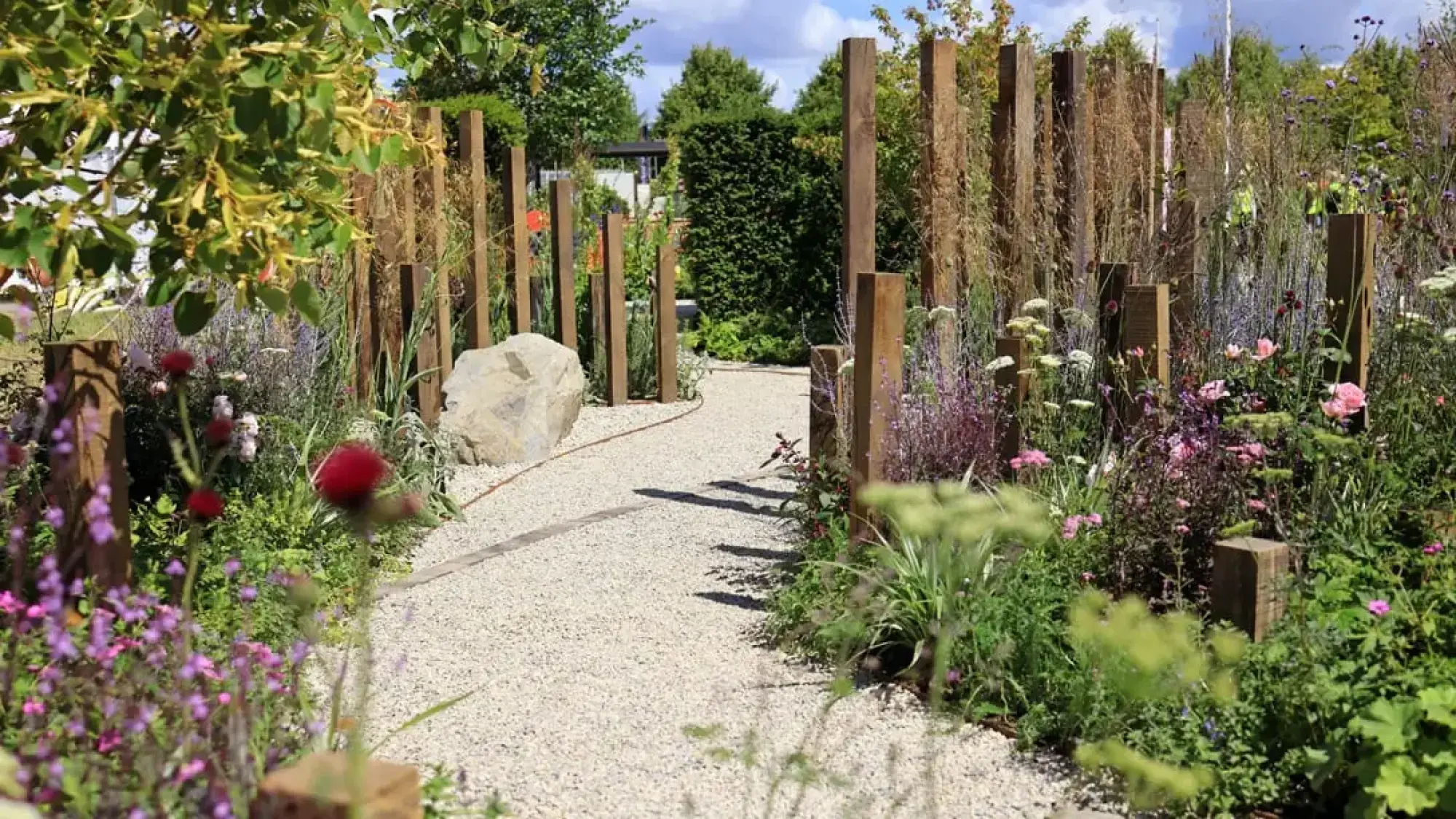 A naturalistic garden features tall timber posts framing a winding gravel path, leading past a large boulder and borders of purple, pink, and white meadow flowers.