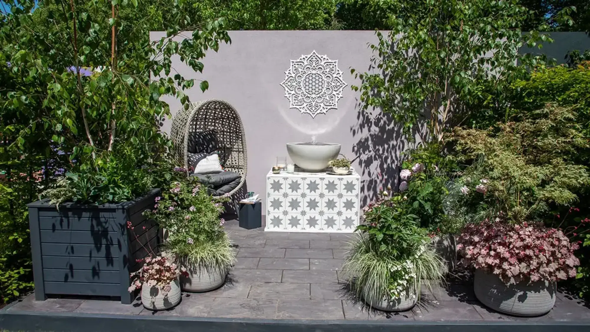A tranquil container garden featuring a white mandala wall art above a water feature, flanked by a woven egg chair and containerised birch trees.