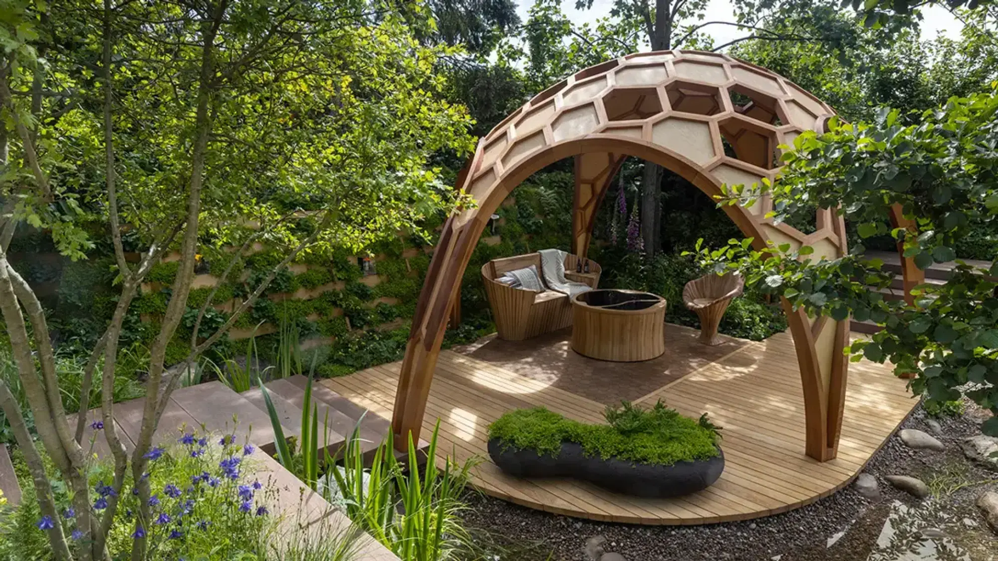 A contemporary garden features a large, honeycomb-patterned wooden pavilion, a curved timber deck with matching seating, and lush green planting.