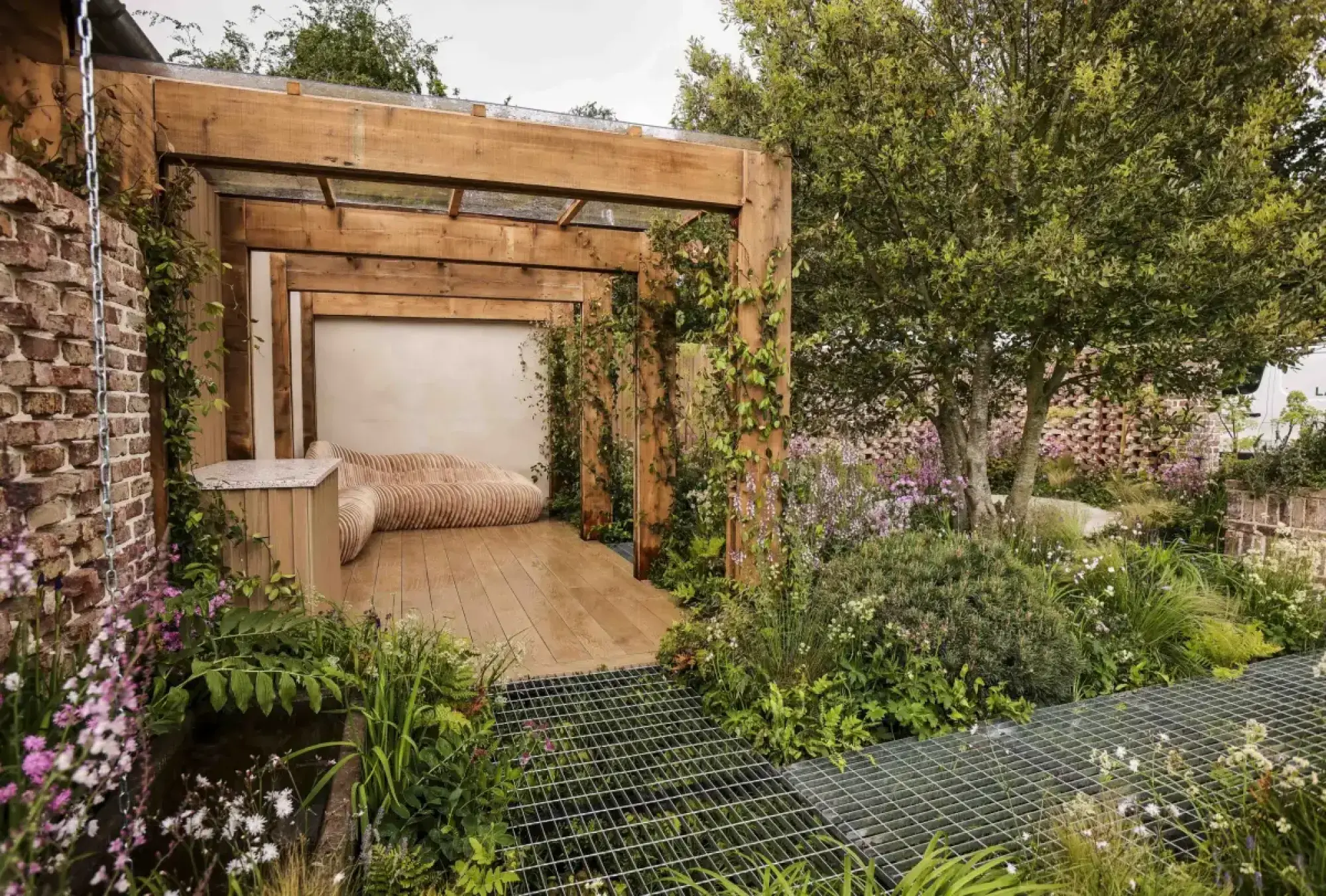 A modern urban garden features a timber pergola with a curved slatted sofa and wooden deck, flanked by a metal grid path through lush planting and a small pond.