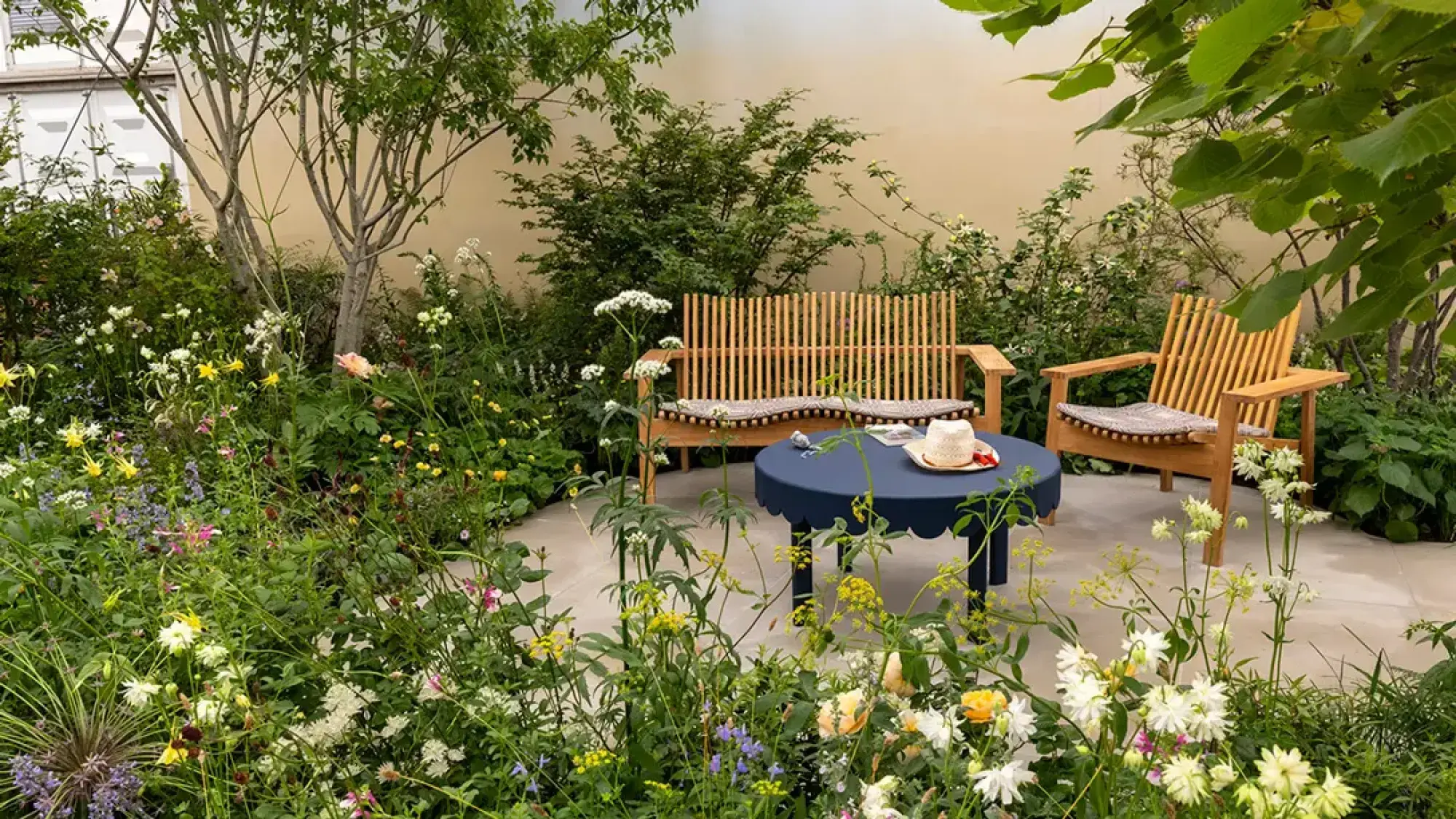 A calm garden with naturalistic planting of mixed flowers surrounds pale paving, wooden slat seating, and a navy scalloped table.