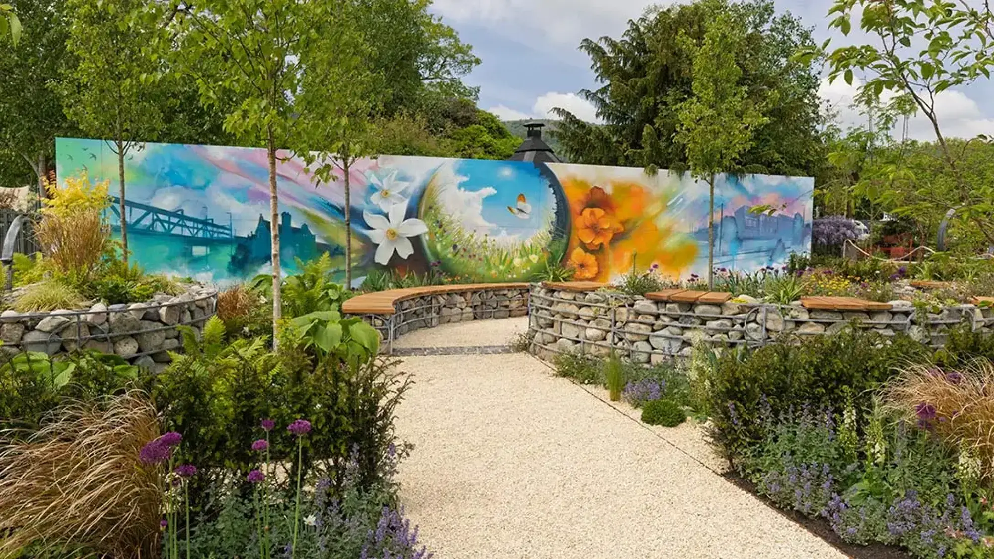 A contemporary urban garden features a vibrant mural on a back wall, gabion basket seating, a gravel path, and mixed herbaceous planting with small trees.