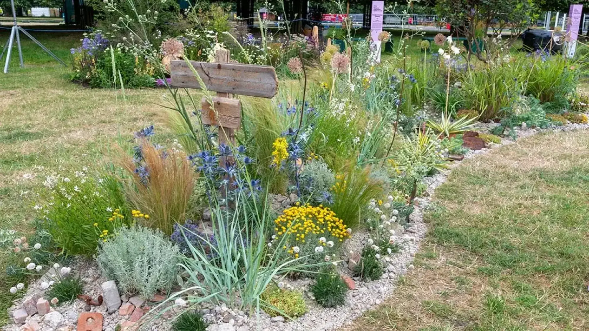 A pocket planting garden features a rustic timber signpost among naturalistic mixed perennials, ornamental grasses, blue sea holly, yellow yarrow, silver foliage, and white flowers, edged with gravel and broken bricks.