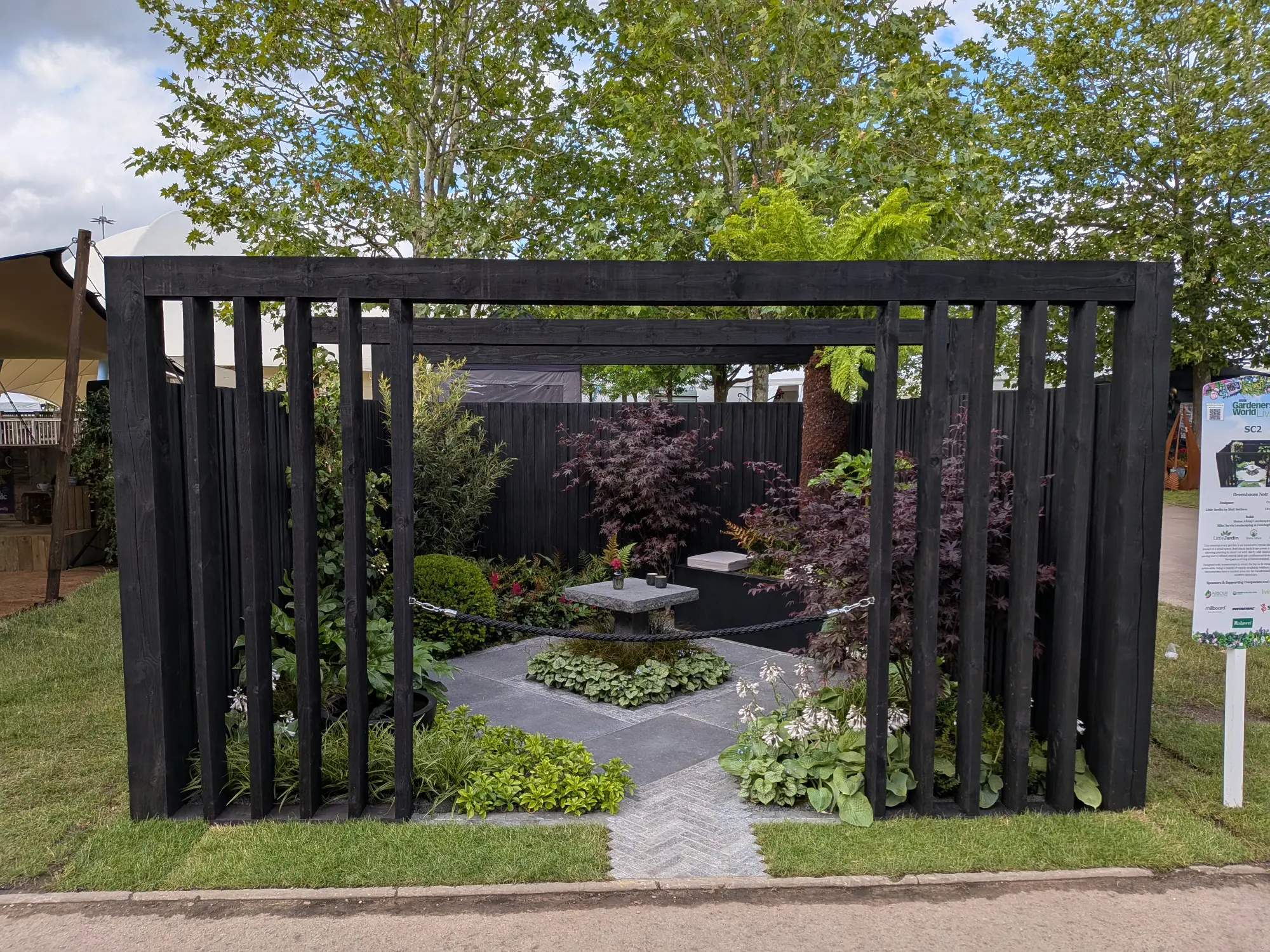 A contemporary showcase garden features a black slatted enclosure, grey paving, a central stone table, and varied low green and dark red foliage plants with white flowers.