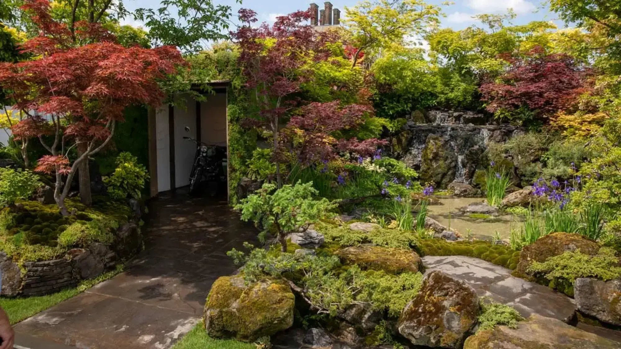A tranquil Japanese sanctuary garden features a waterfall cascading into a pond, a dark stone path, and red Japanese maples beside a timber shelter.