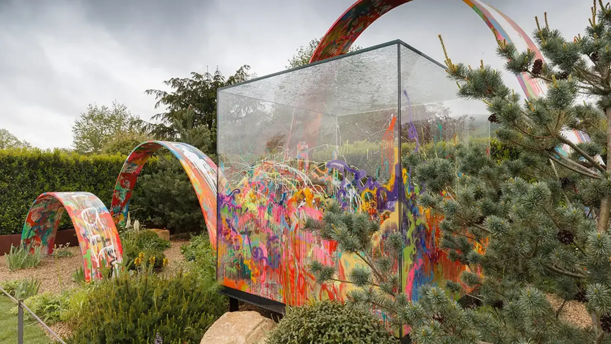A modern show garden featuring a large clear glass cube filled with abstract, multicoloured paint splatters, surrounded by painted arch structures and pine trees.