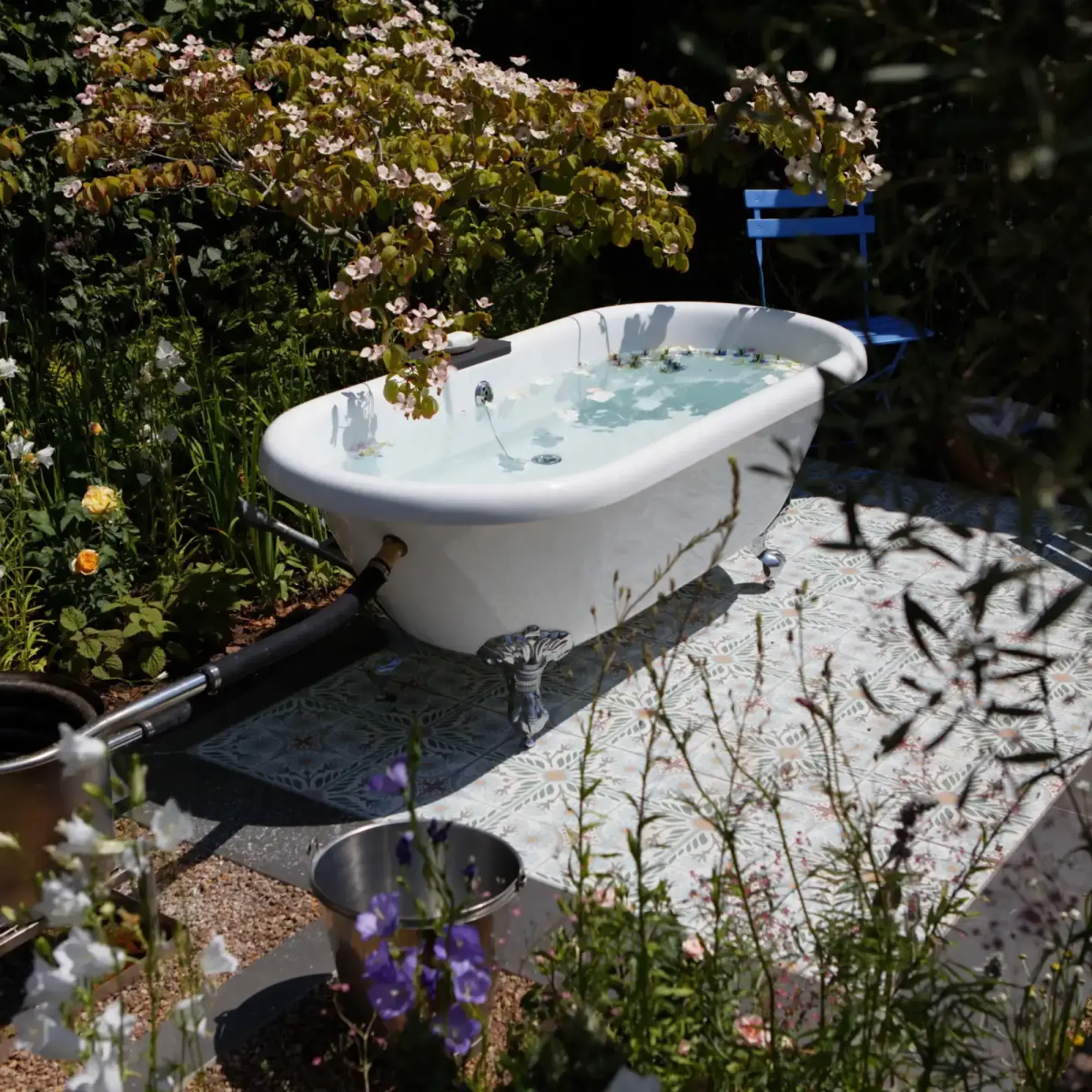 A white clawfoot bathtub filled with water and petals sits on a patterned tiled patio, surrounded by lush green foliage and various flowering plants.