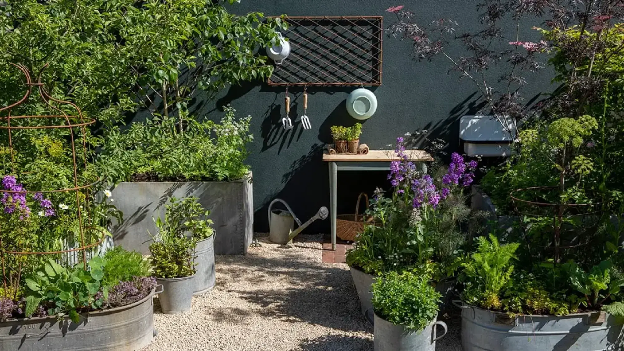 A charming container kitchen garden featuring various herbs and edible plants in galvanized tubs and pots, set against a dark grey wall with a rustic potting bench.