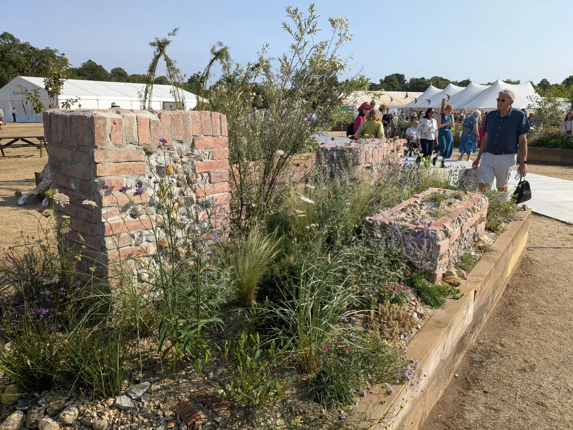 A long border with reclaimed brick and flint structures, timber edging, and a mix of drought-tolerant grasses, purple flowers, and low shrubs.