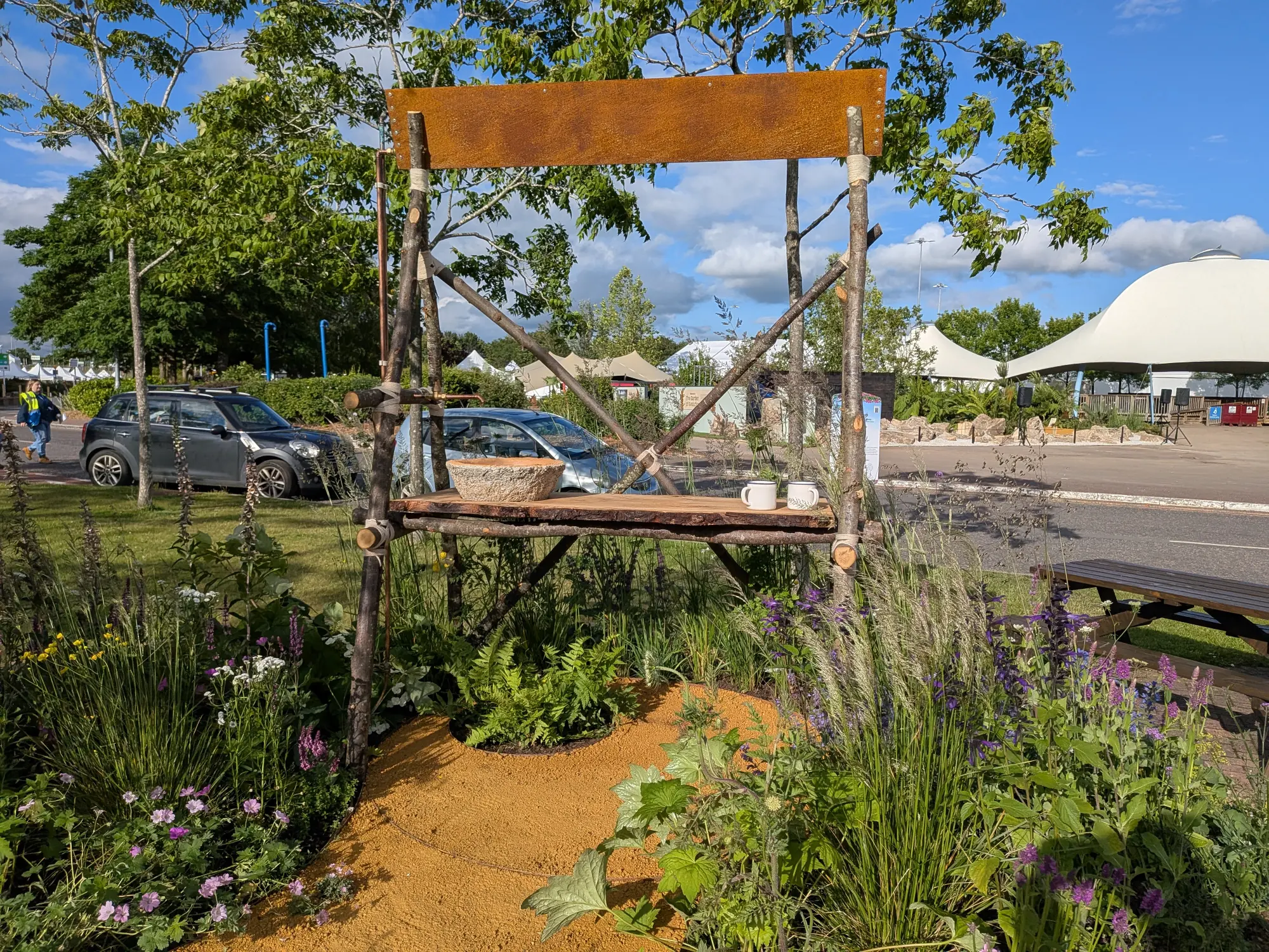 A beautiful border with a rustic timber A-frame, rusted metal sign, shelf, and an ochre path through grasses, purple flowers, and ferns.