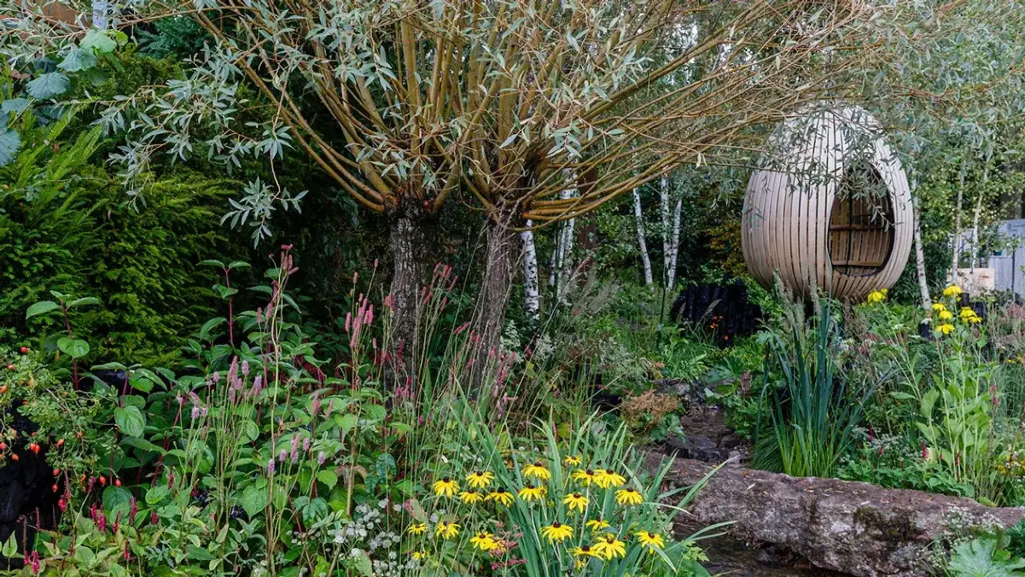 An immersive organic show garden featuring an egg-shaped wooden pod, yellow coneflowers, pink spires, a multi-stemmed willow, and lush mixed planting.