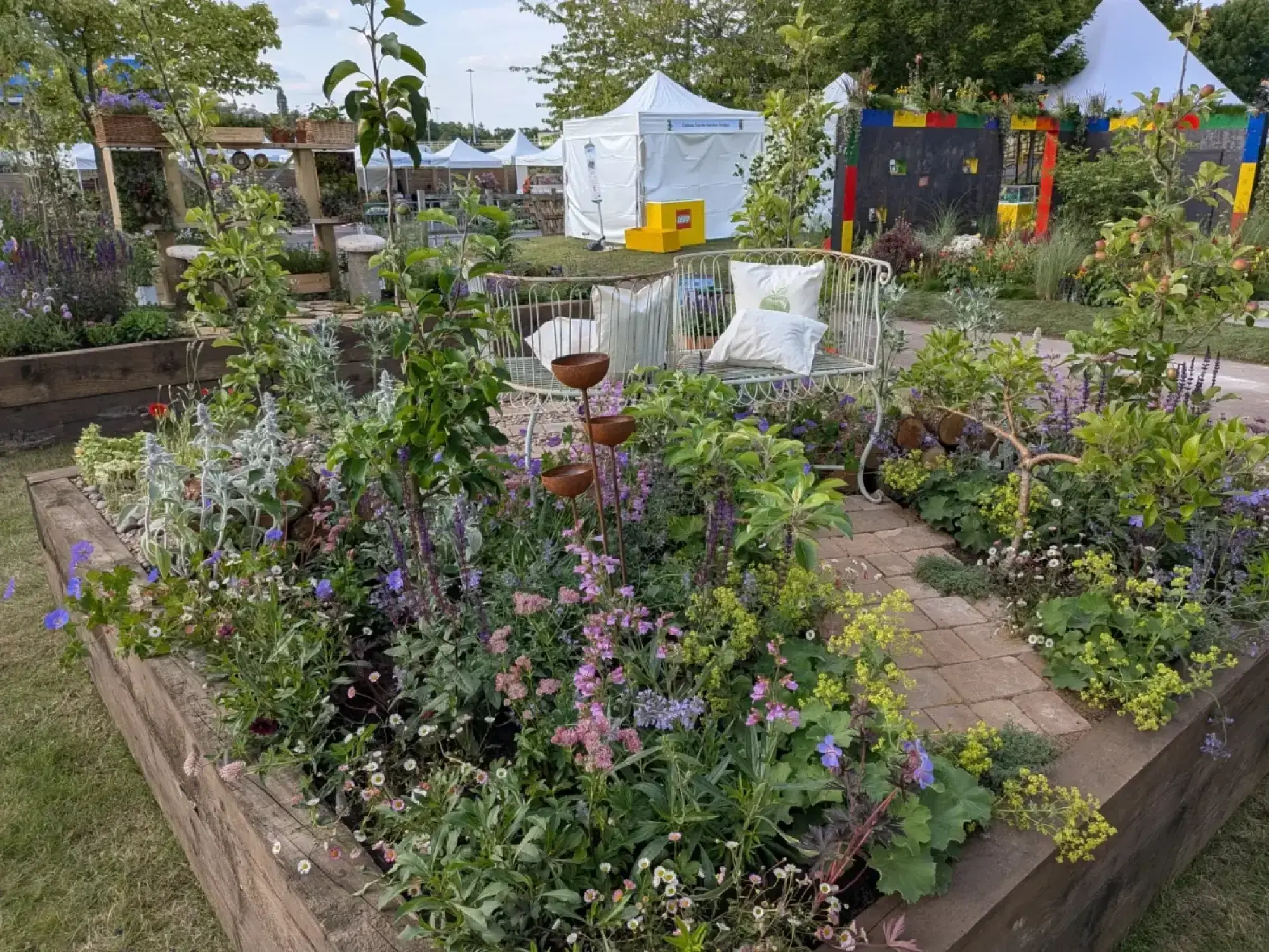 A raised timber border garden showcases young apple trees, a white metal bench, a tiered rusty water feature, a brick path, and vibrant perennial flowers.