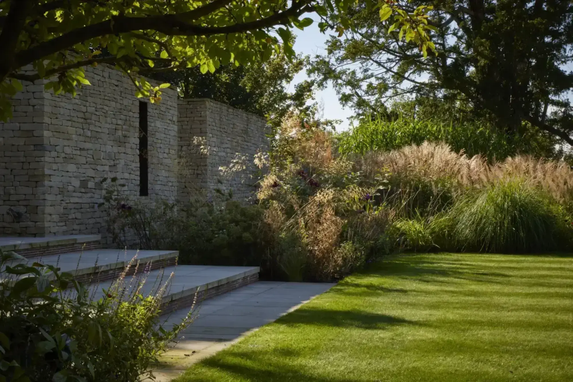 A formal Arts and Crafts garden featuring terraced dry stone walls, wide flagstone steps, a lush green lawn, and abundant ornamental grasses.