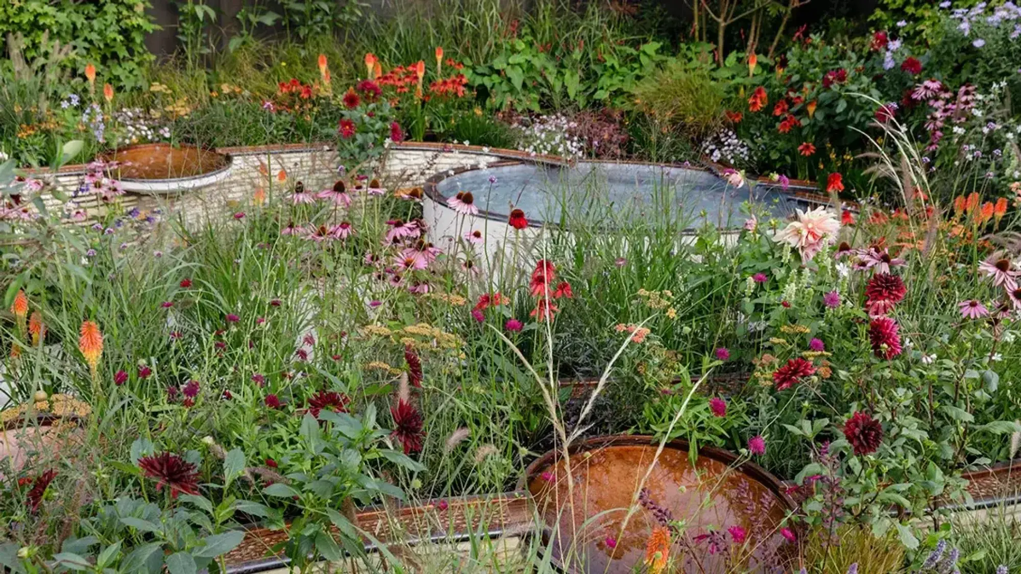 A vibrant sanctuary garden features curved, mosaic-tiled walls with copper water bowls, a circular pool, and lush, naturalistic planting of pink echinacea, red hot pokers, and burgundy dahlias amidst tall grasses.