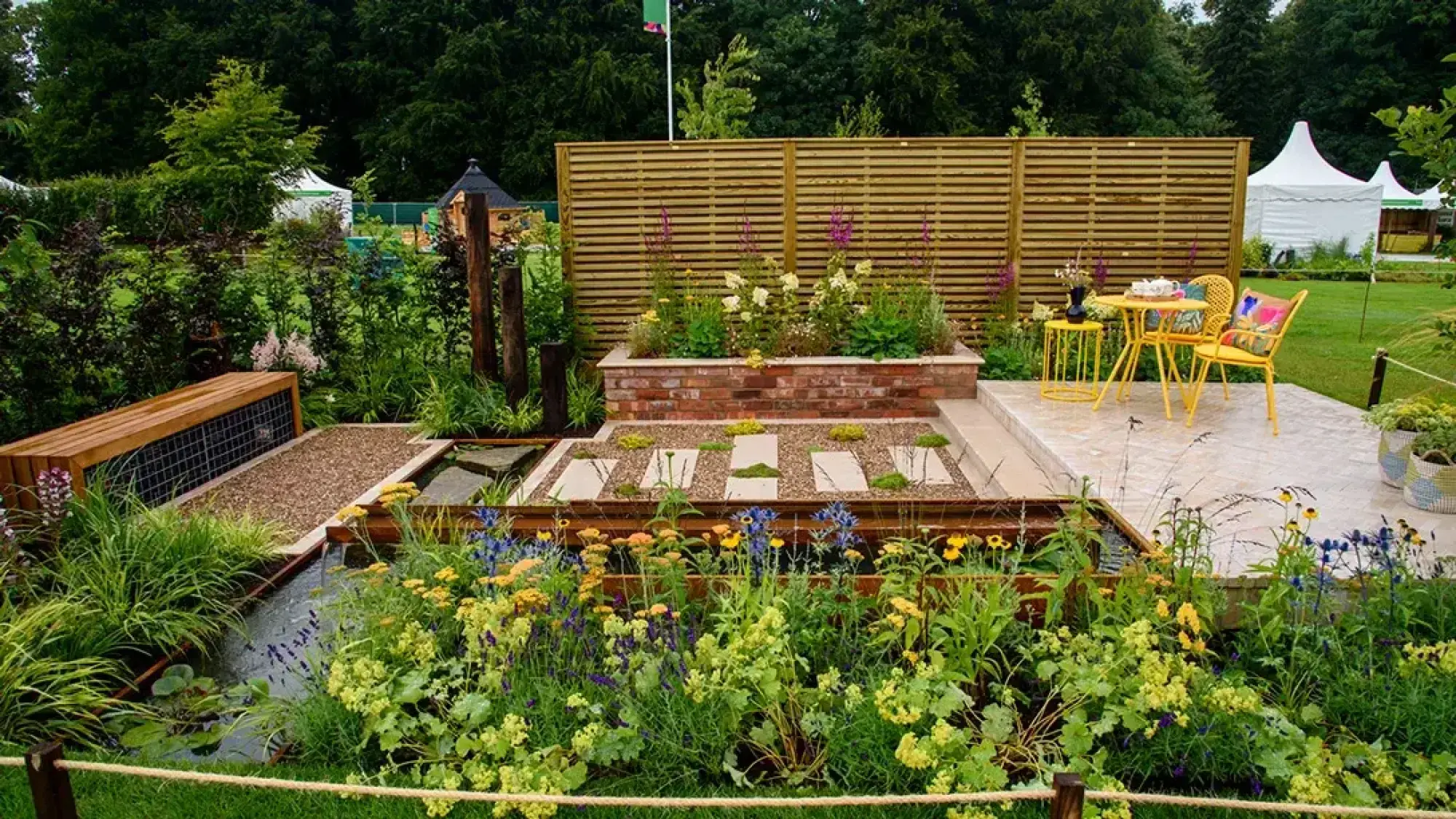 A contemporary garden with a gabion timber bench, multi-level water feature, brick raised bed, and a patio with yellow bistro furniture, surrounded by lush mixed planting.