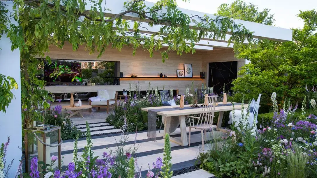 A modern outdoor room with an outdoor television, dining and lounge areas under a pergola, surrounded by purple and white flowers.