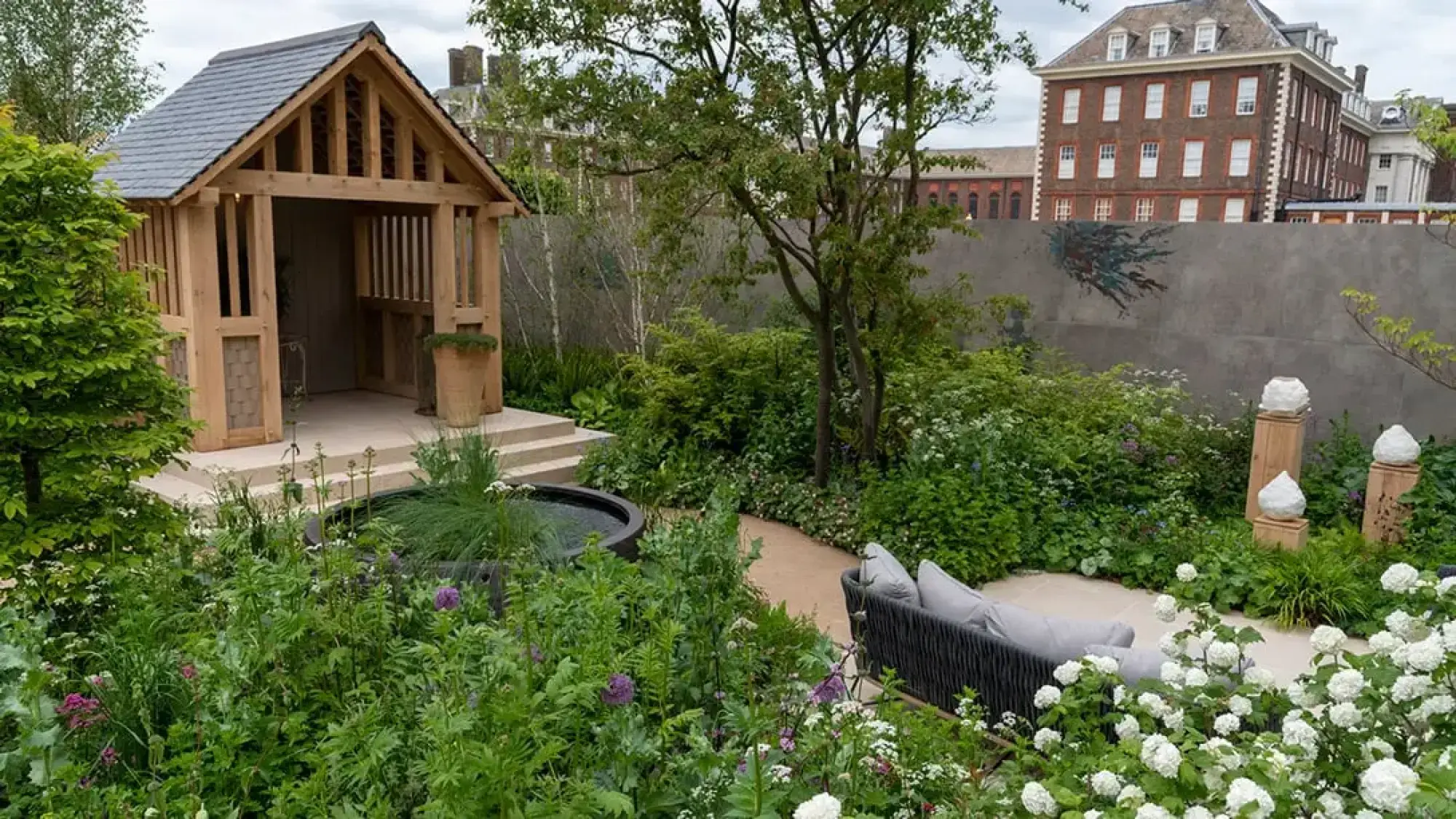 A serene show garden featuring a timber pavilion, circular water feature, woven sofa, and lush perennial planting with white and purple flowers.