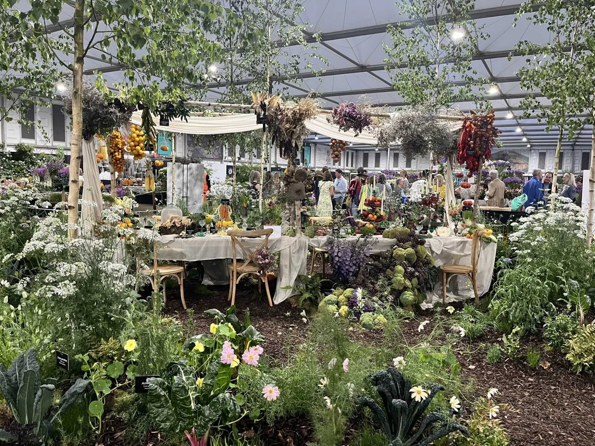 The 'Feast' garden features a long white dining table amidst edible plants and flowers, with colourful hanging produce and birch trees.