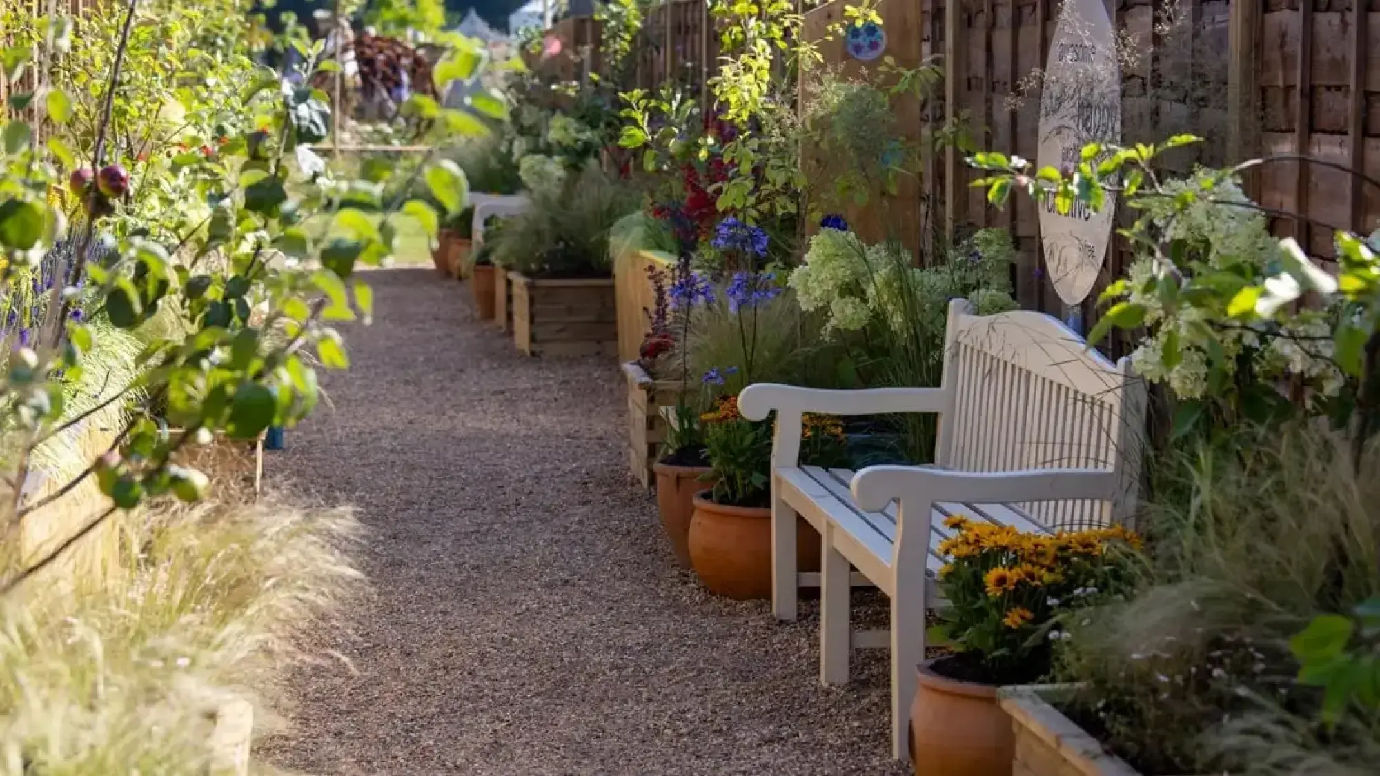 A narrow ginnel garden features a gravel path bordered by timber planters and terracotta pots with vibrant mixed planting and a white wooden bench.