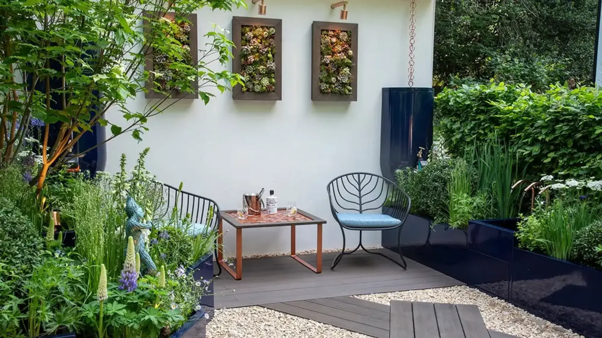 A modern container garden has vertical succulent planters, blue trough planters with lush greenery, and a seating area on grey decking.