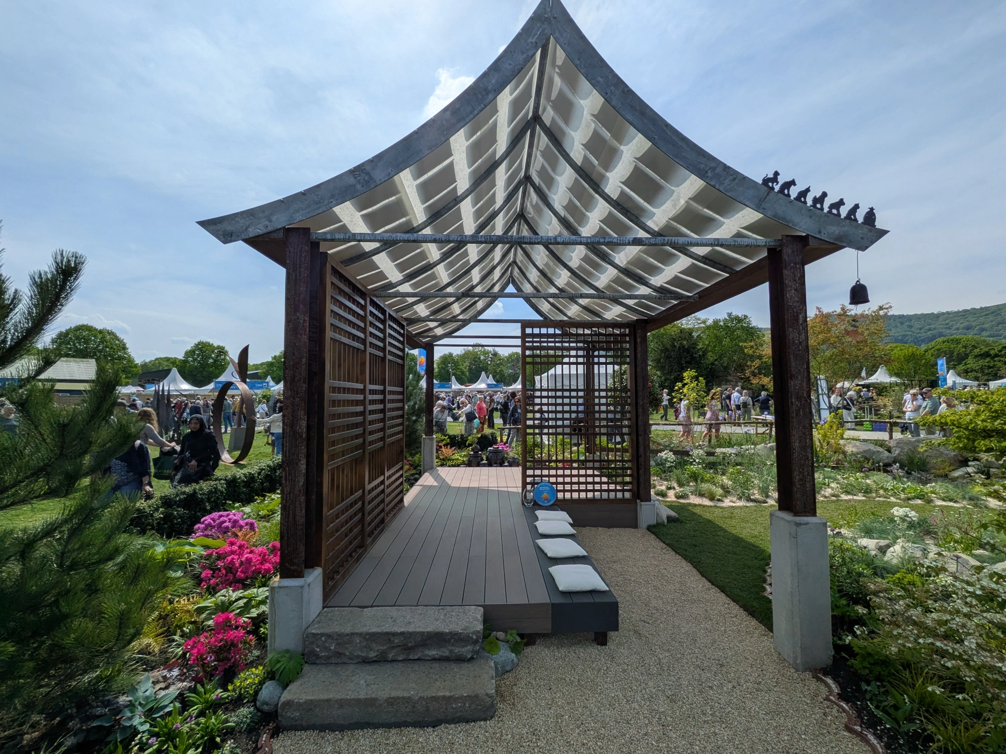 A serene Asian-inspired garden with a curved-roof pavilion, slatted timber walls, a composite deck, stone steps, and vibrant pink azaleas.