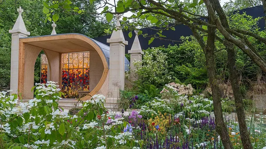 A show garden featuring a curved timber pavilion with a vibrant red-orange stained glass window, flanked by stone pillars and lush borders of white, purple, yellow, and blue flowering plants.