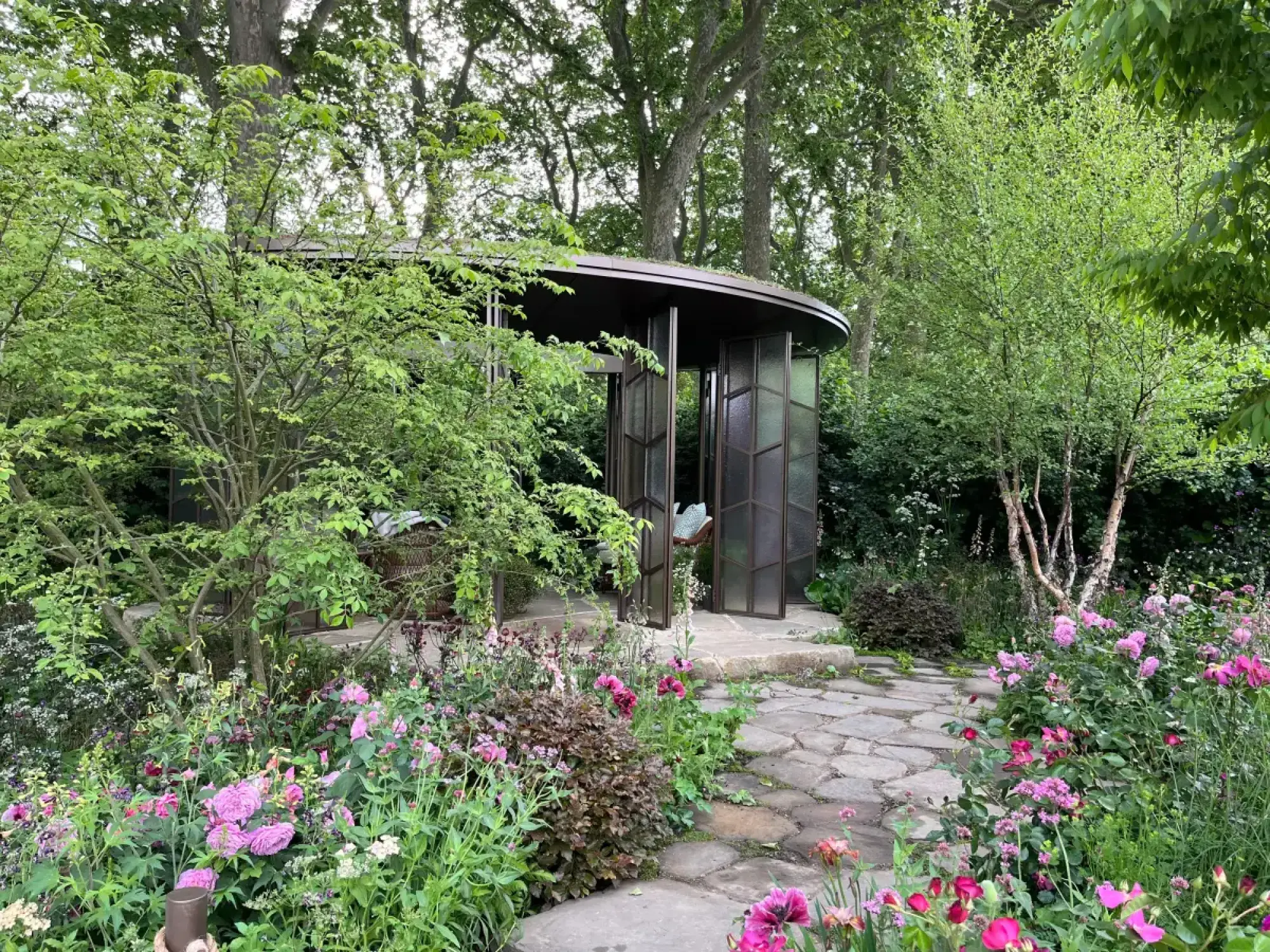 A curved glasshouse with a green roof sits in a naturalistic garden with a flagstone path, pink roses, purple flowers, and white birch trees.