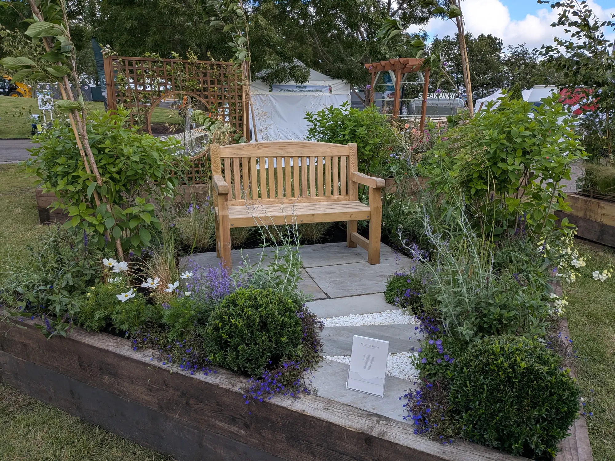 A tranquil border garden features a wooden bench centered on grey flagstone paving and white gravel, surrounded by lush green shrubs, purple lobelia, and white cosmos flowers.
