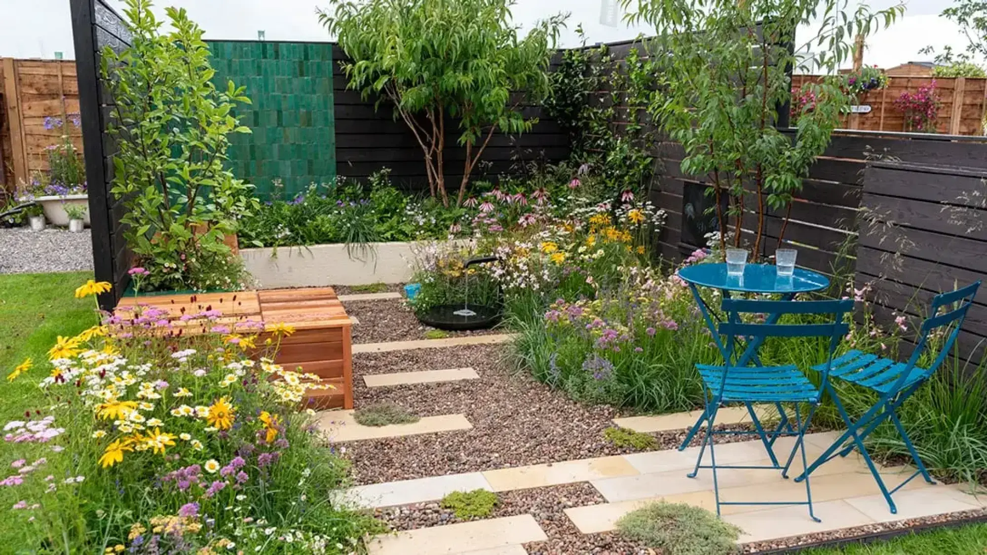 A vibrant terrace garden with a teal bistro set and timber bench, featuring a water feature and colourful wildflower planting.