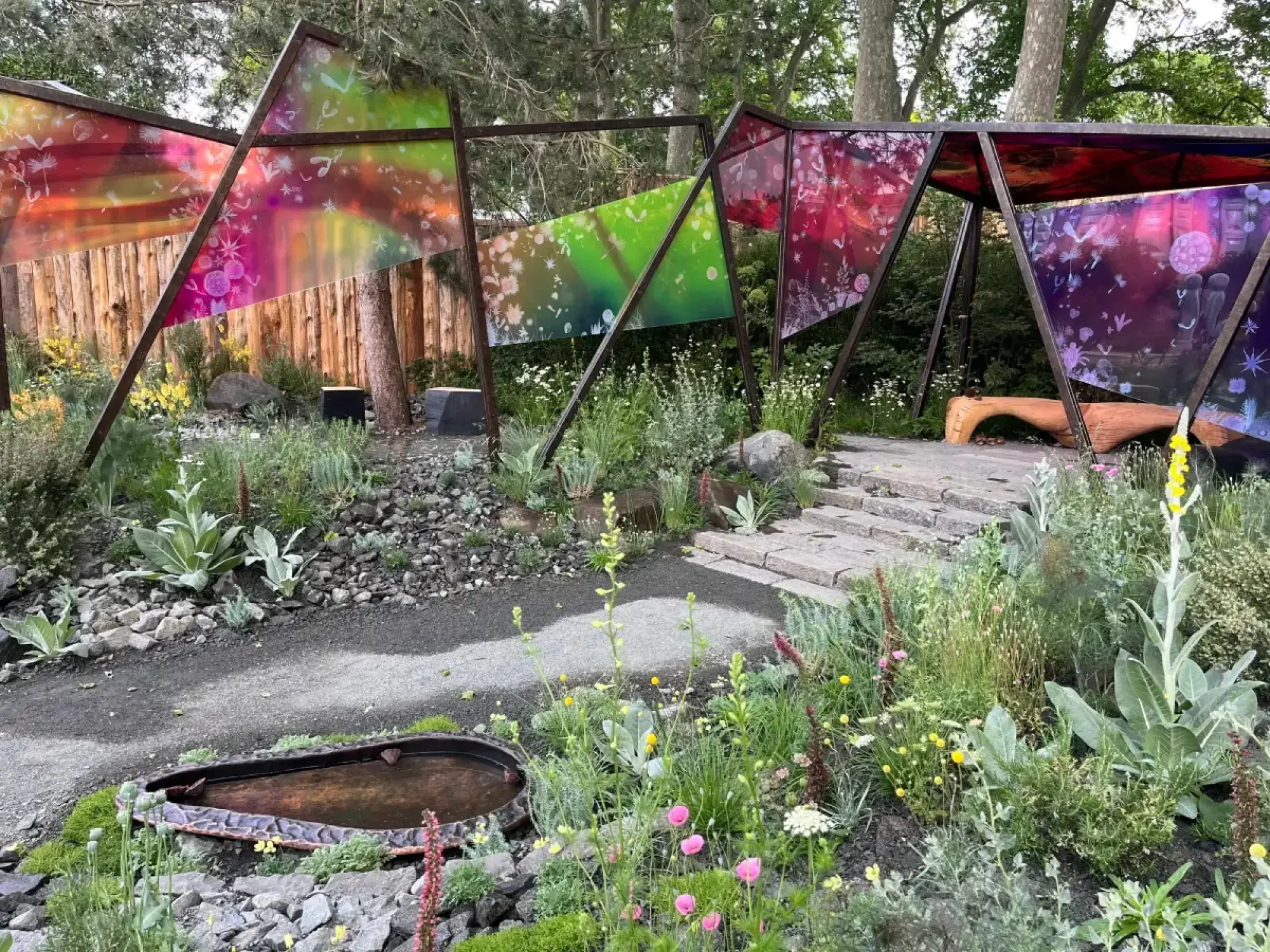 A contemporary show garden features a geometric pavilion with vibrant, patterned translucent panels, log seating, naturalistic planting, and a water trough.