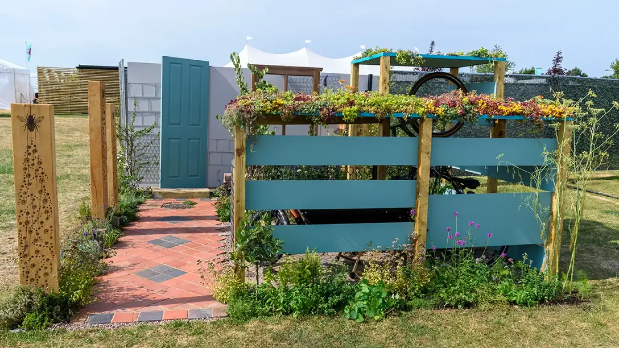 A greener front garden features a teal bike shelter, sedum roof, bicycle wheel, brick path, bee-carved posts, and pollinator plants.