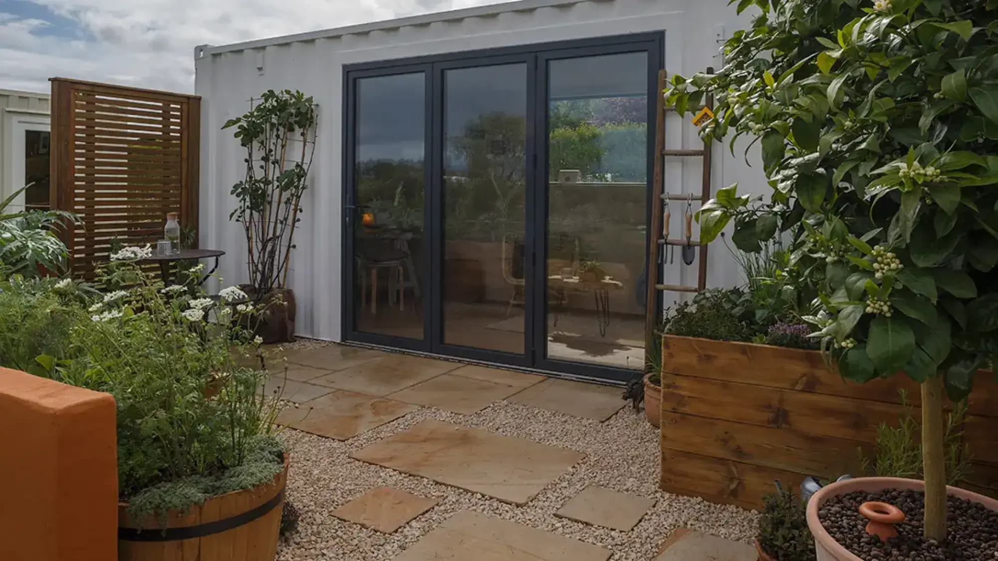 A modern urban patio garden featuring a white shipping container home with dark bifold doors, surrounded by wooden planters containing edible plants, herbs, and a citrus tree.