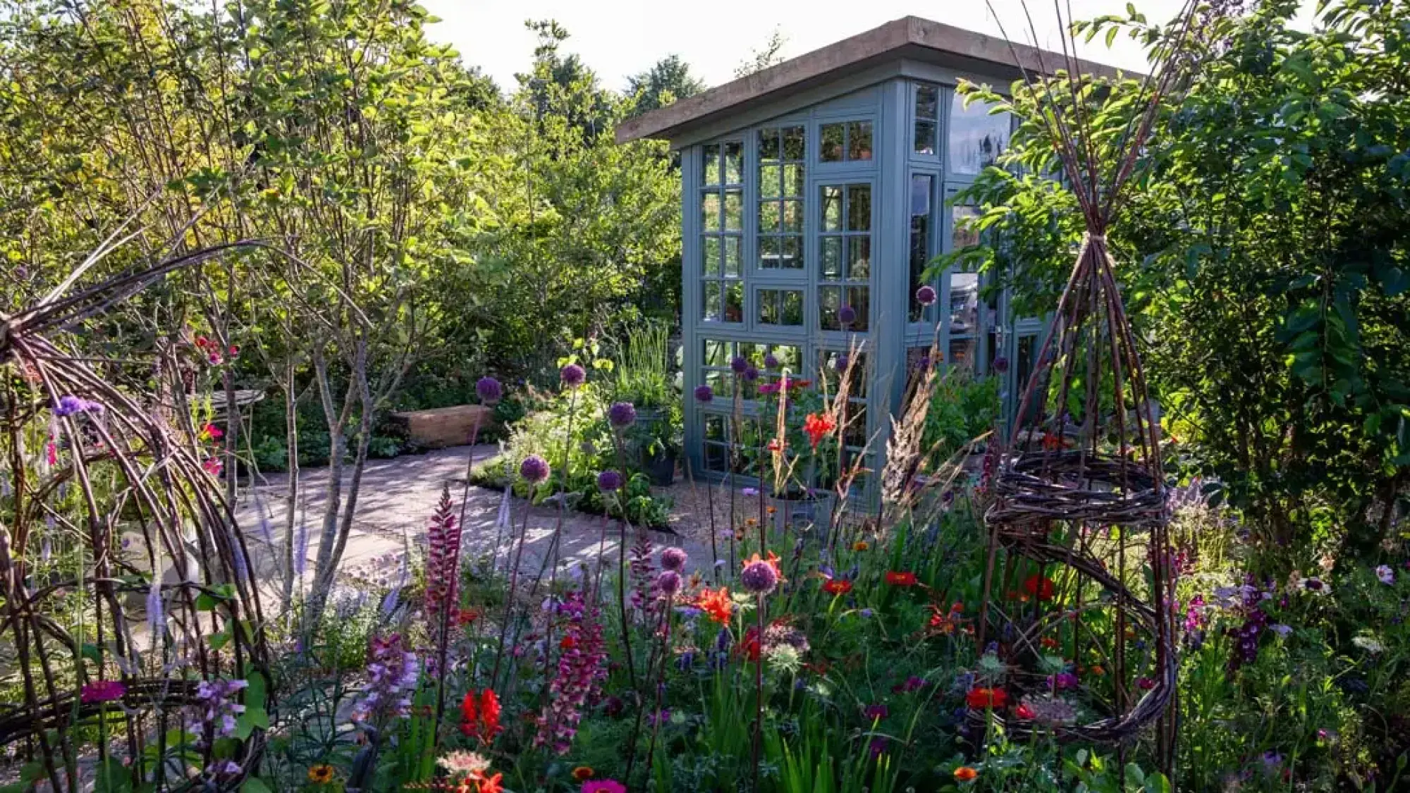 A vibrant, informal garden features a green greenhouse with many windows, a light gravel path, and colourful mixed planting with purple alliums and woven willow structures.