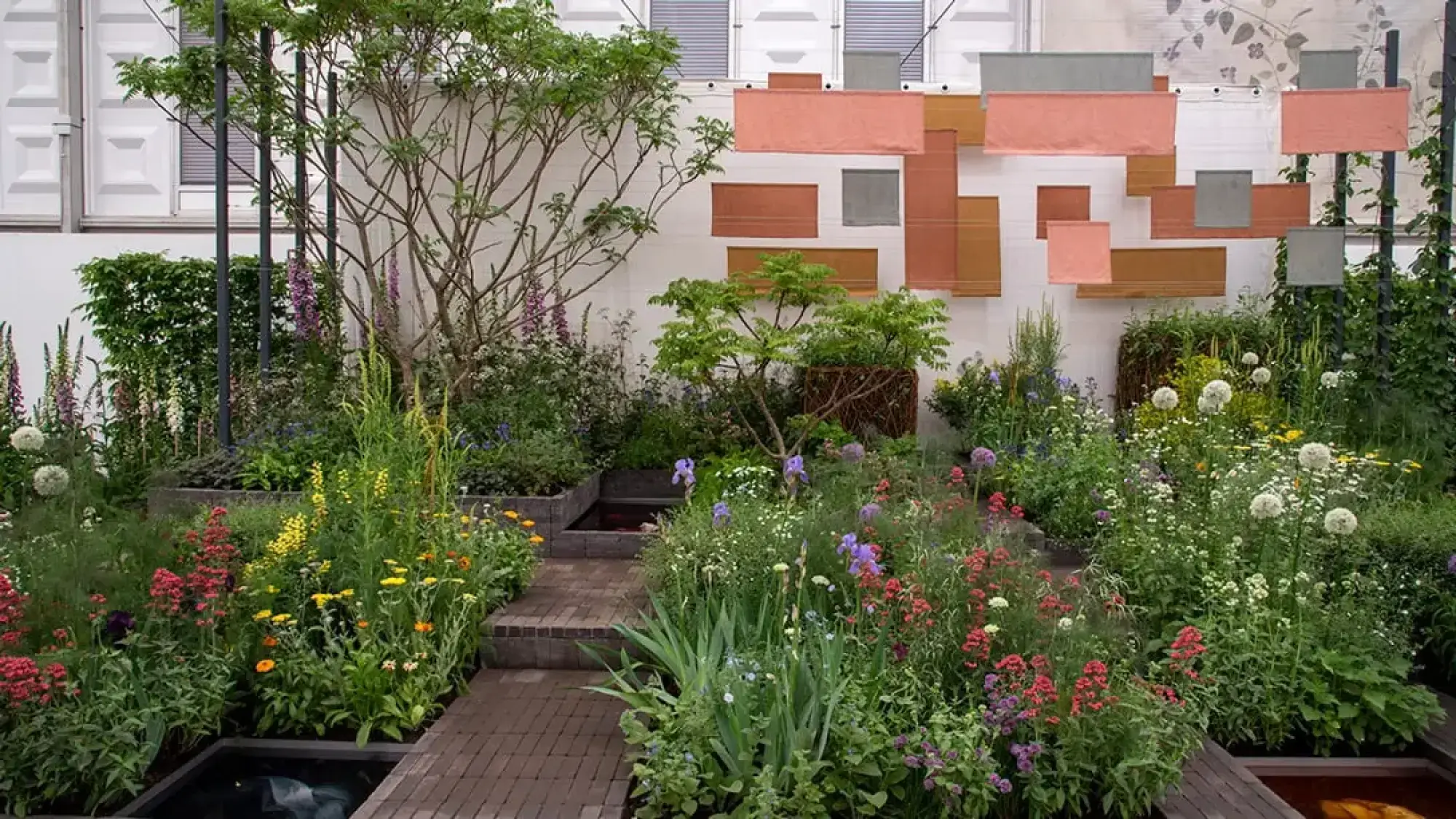 A colourful garden with a brick path, water channels, and varied flowers, backed by a white wall displaying floating terracotta and grey fabric panels.