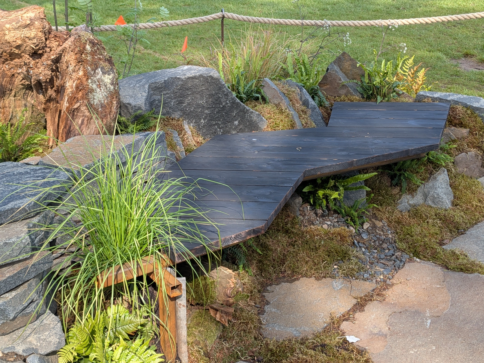 A naturalistic border garden with a dark charred timber walkway, large granite boulders, moss, ferns, and an ancient tree stump.