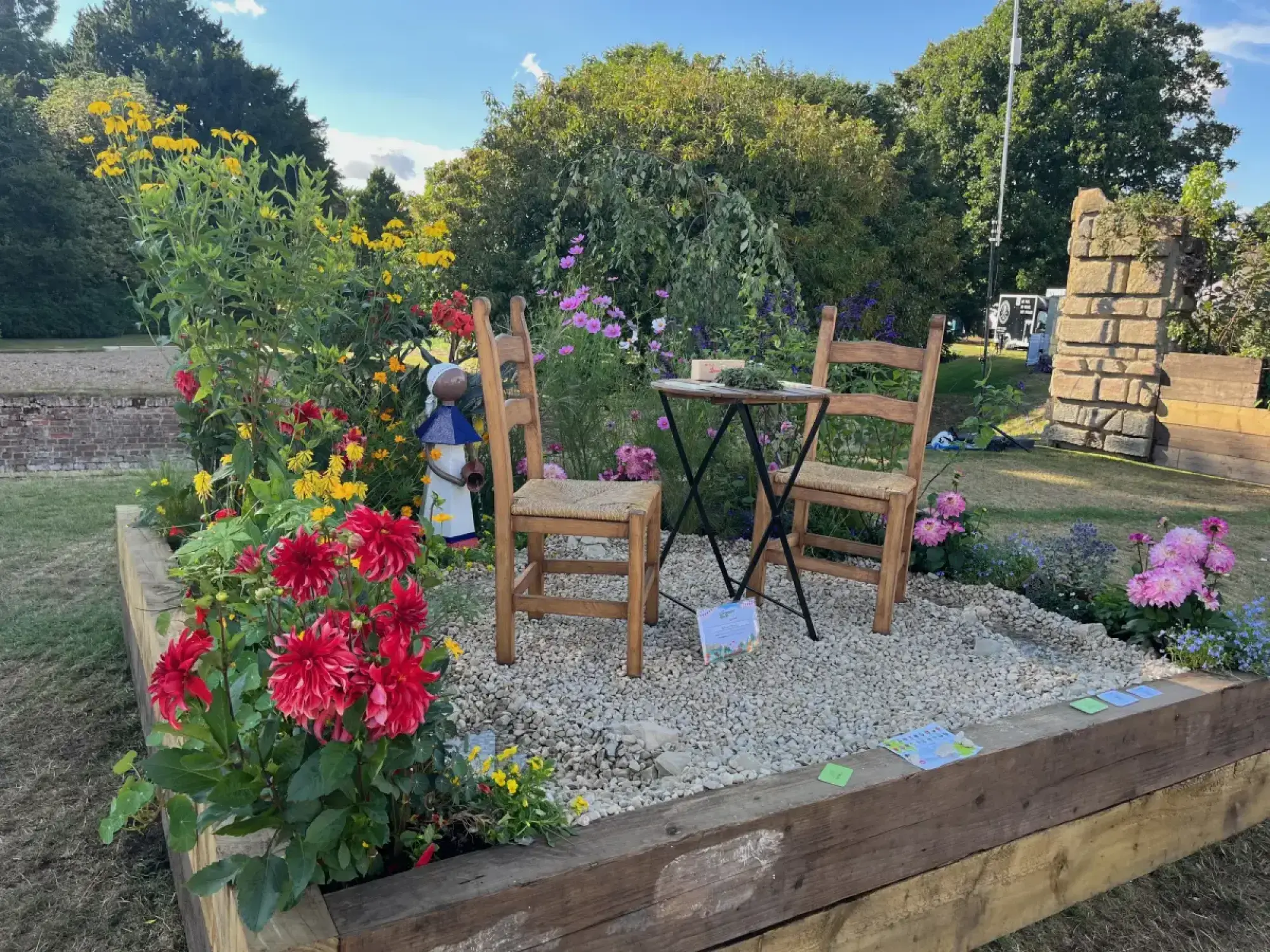 A colourful raised border features a metal figure, bistro table with wooden chairs on gravel, and vibrant red, yellow, pink, and blue flowers.